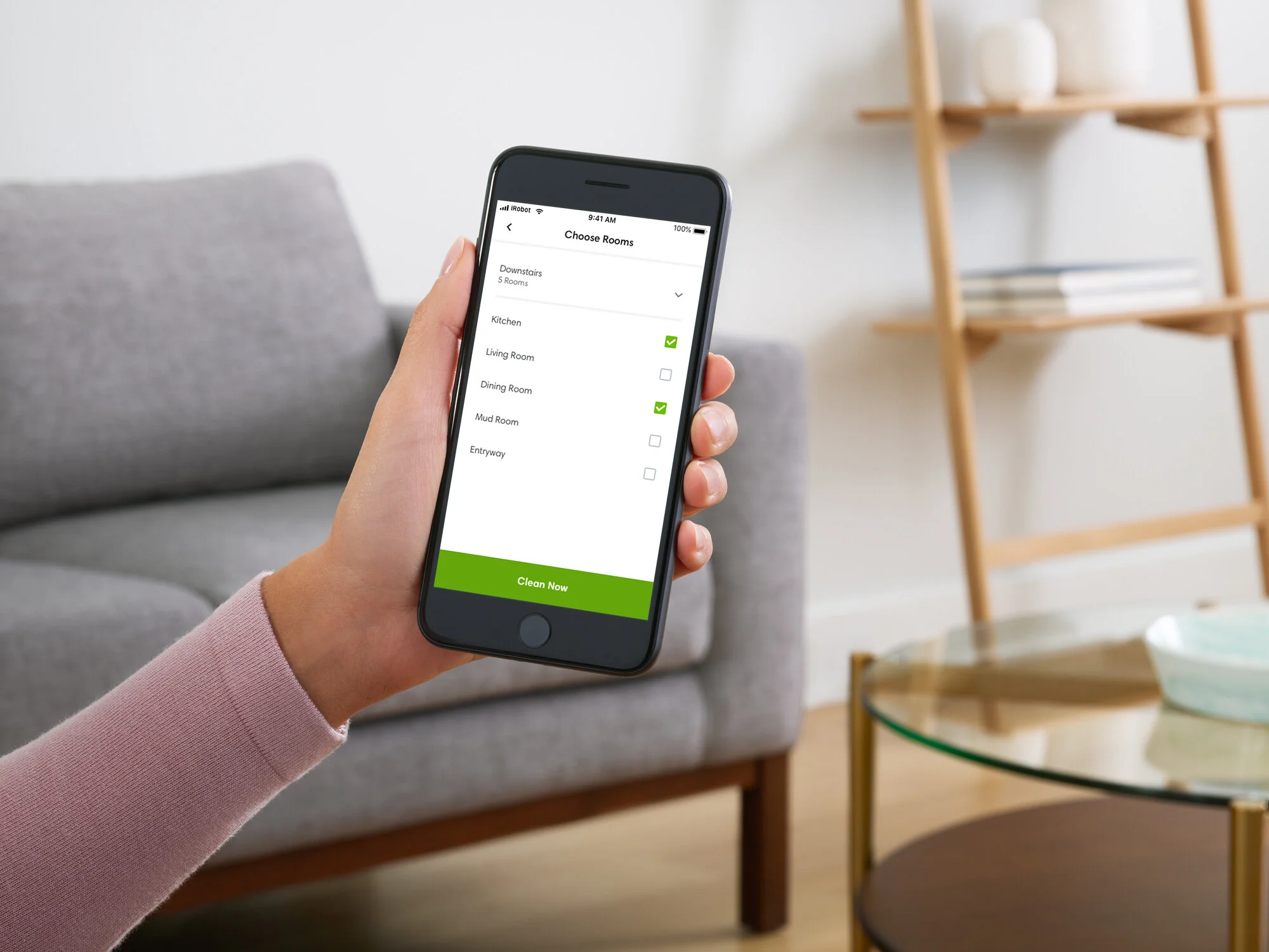 Person holding smartphone with a smart home app for selecting rooms, showing options for kitchen and mud room selected, in a living room with a gray sofa and wooden shelves. Photographed by Joseph Keller for iRobot