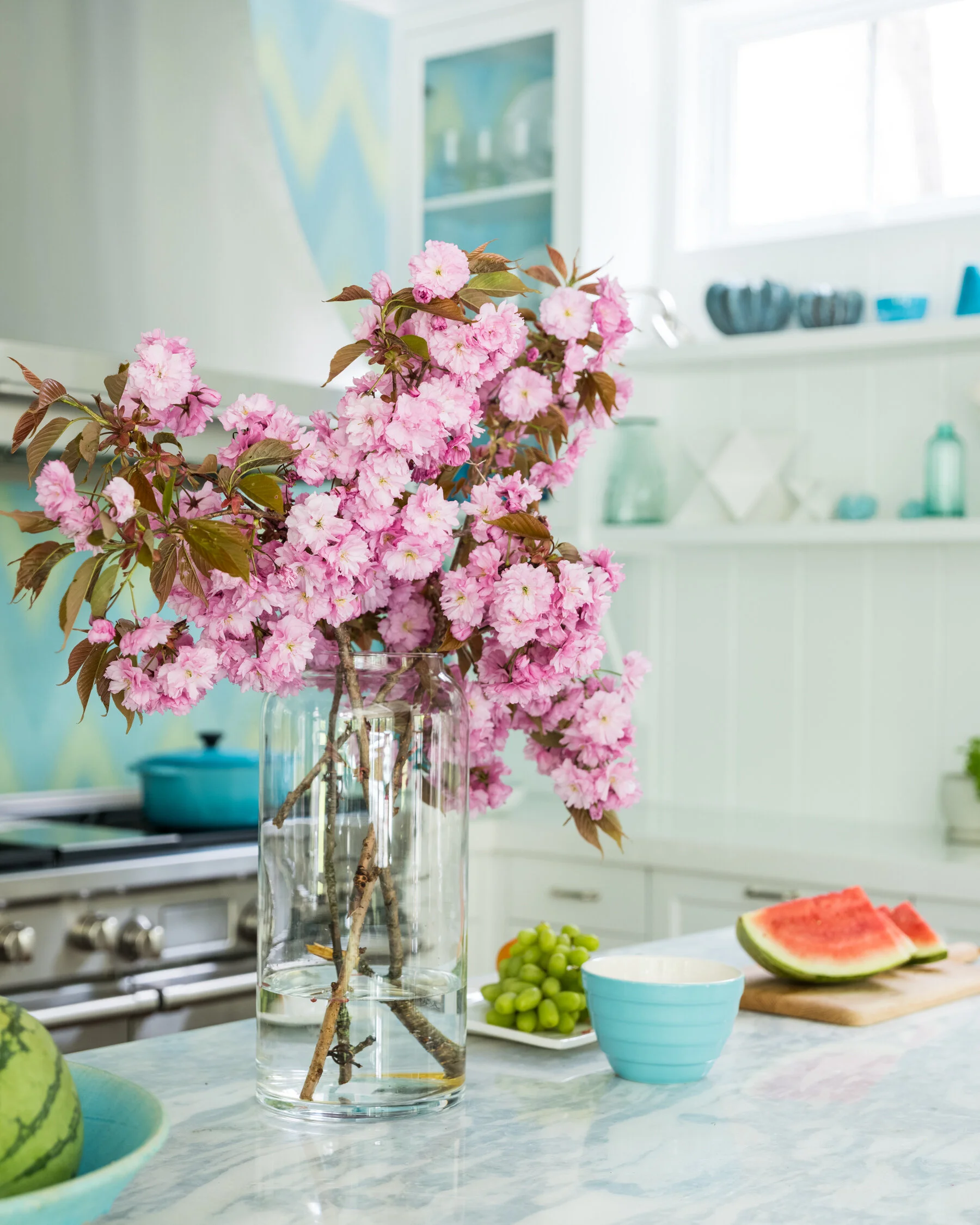 Pink cherry blossom branches in a glass vase on a kitchen countertop with watermelon, grapes, and a bowl nearby
