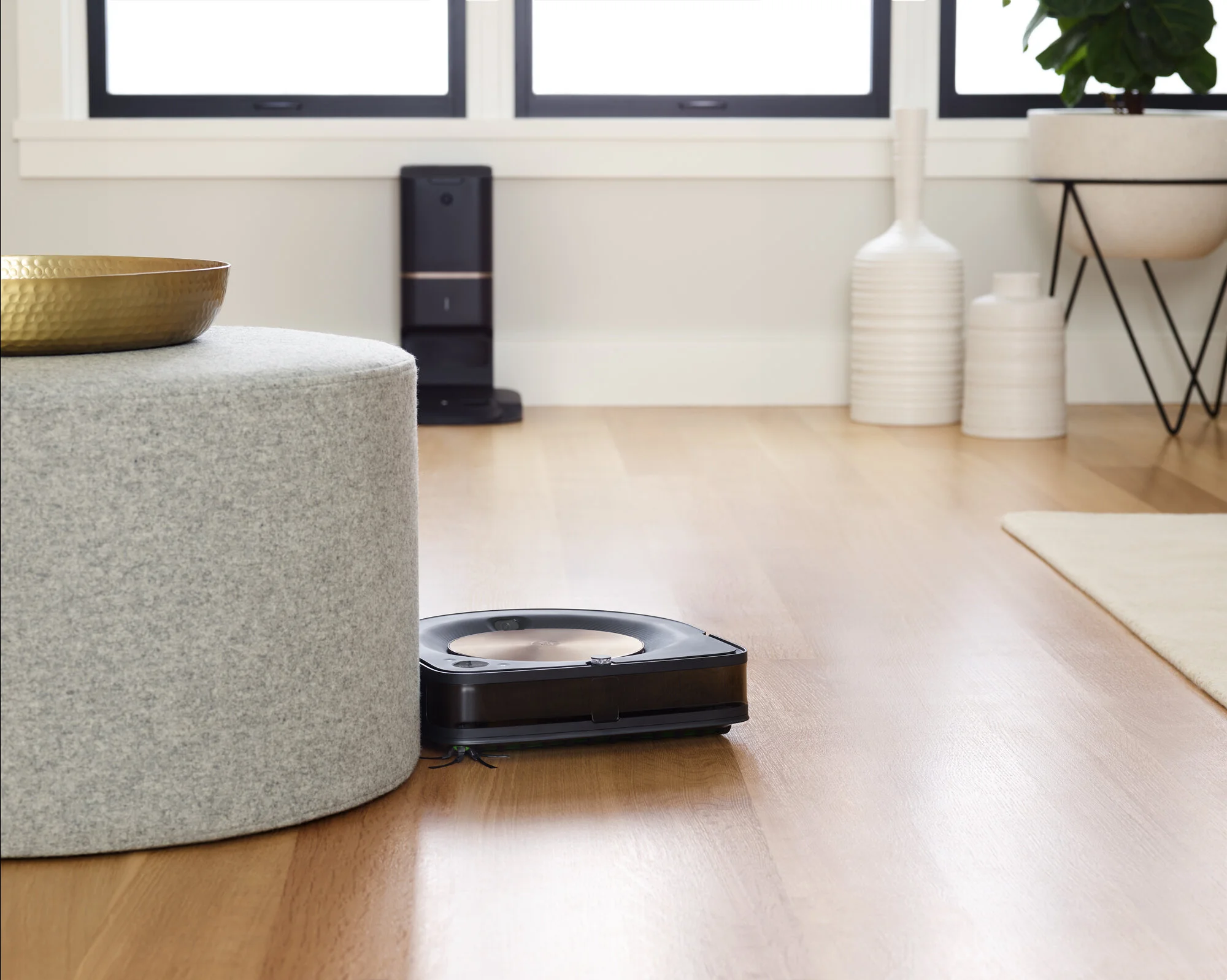 A robotic vacuum cleaner on a wooden floor in a modern living room with a gray upholstered ottoman, a gold bowl on top, and various white decorative vases and planters near a window. Photographed by Joseph Keller for iRobot