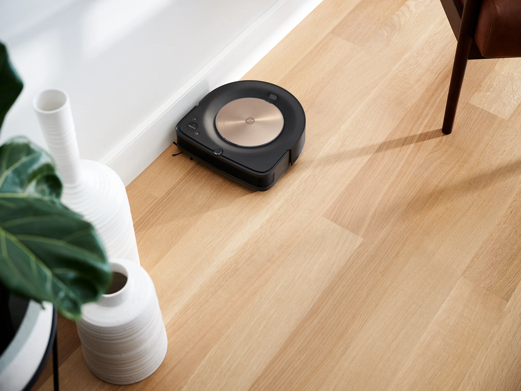 A robotic vacuum cleaner on a wooden floor near a white vase with a green plant, a white striped vase, and a wooden chair leg. Photographed by Joseph Keller for iRobot
