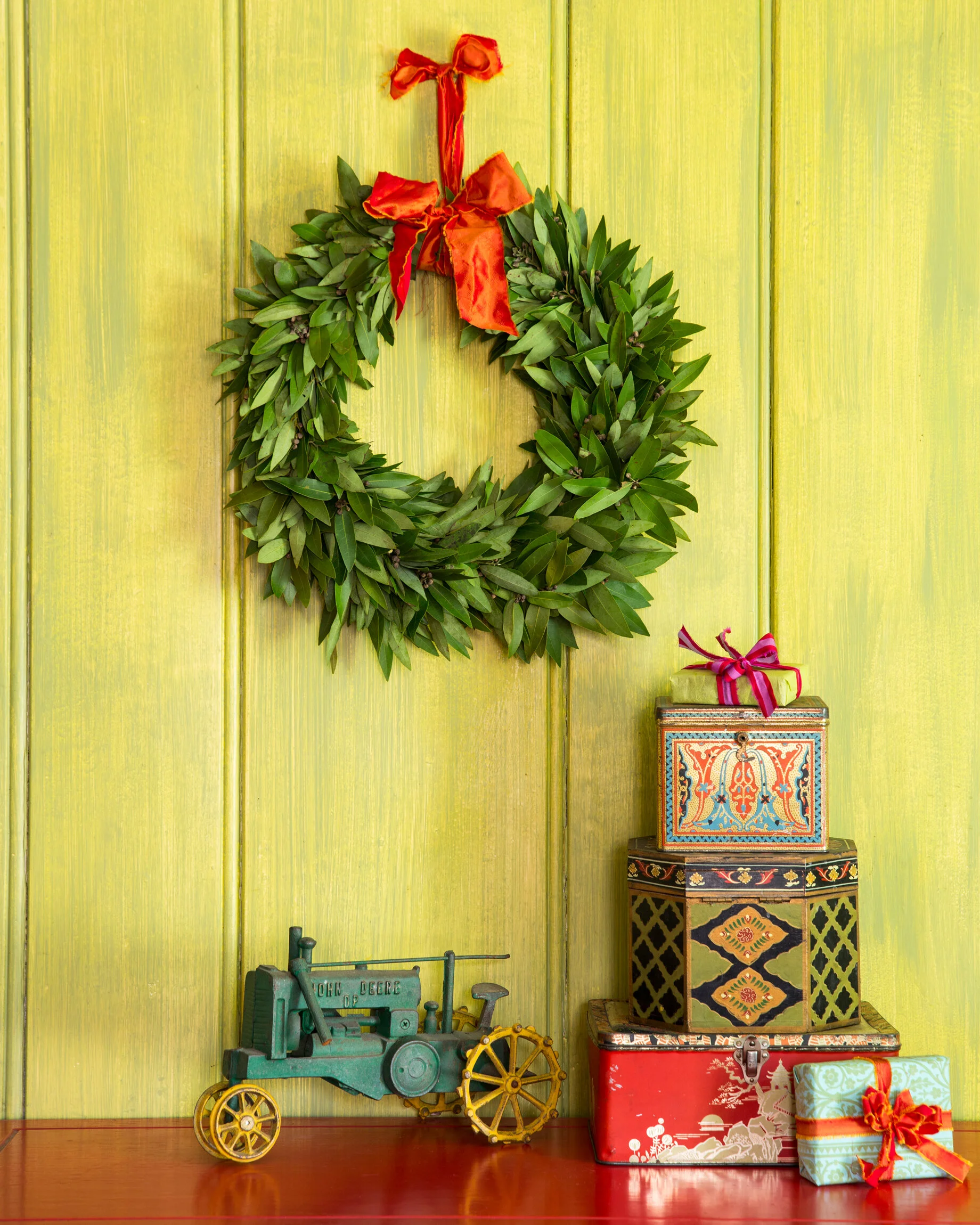 Holiday decor with a green wreath on a yellow wooden wall, decorated with a red ribbon, and three wrapped gifts stacked on decorative boxes, next to a vintage toy tractor.