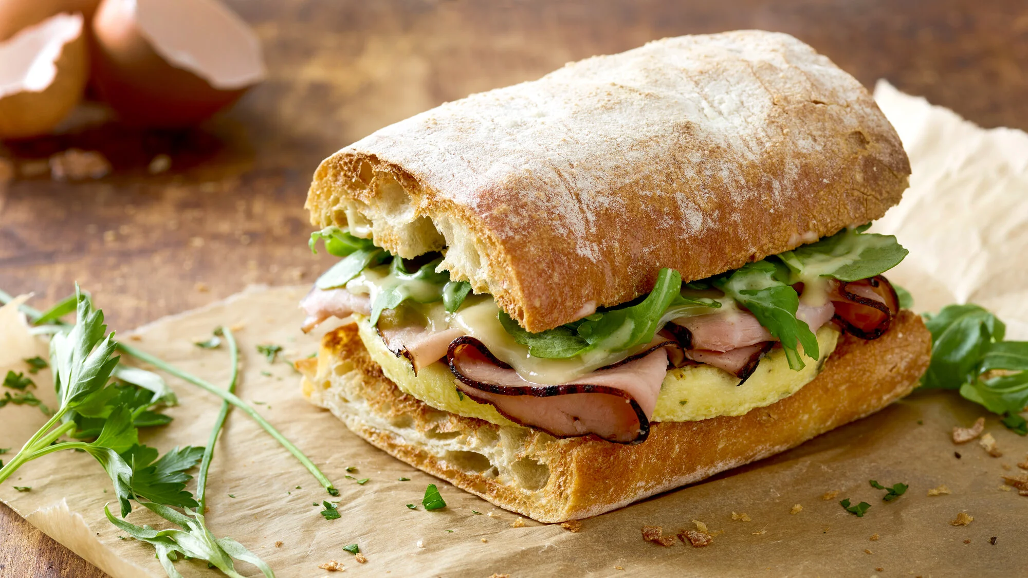 Sandwich on a rustic wooden surface with arugula leaves, featuring ham, cheese, lettuce, and a crusty baguette. Boston based food and hospitality photographer.