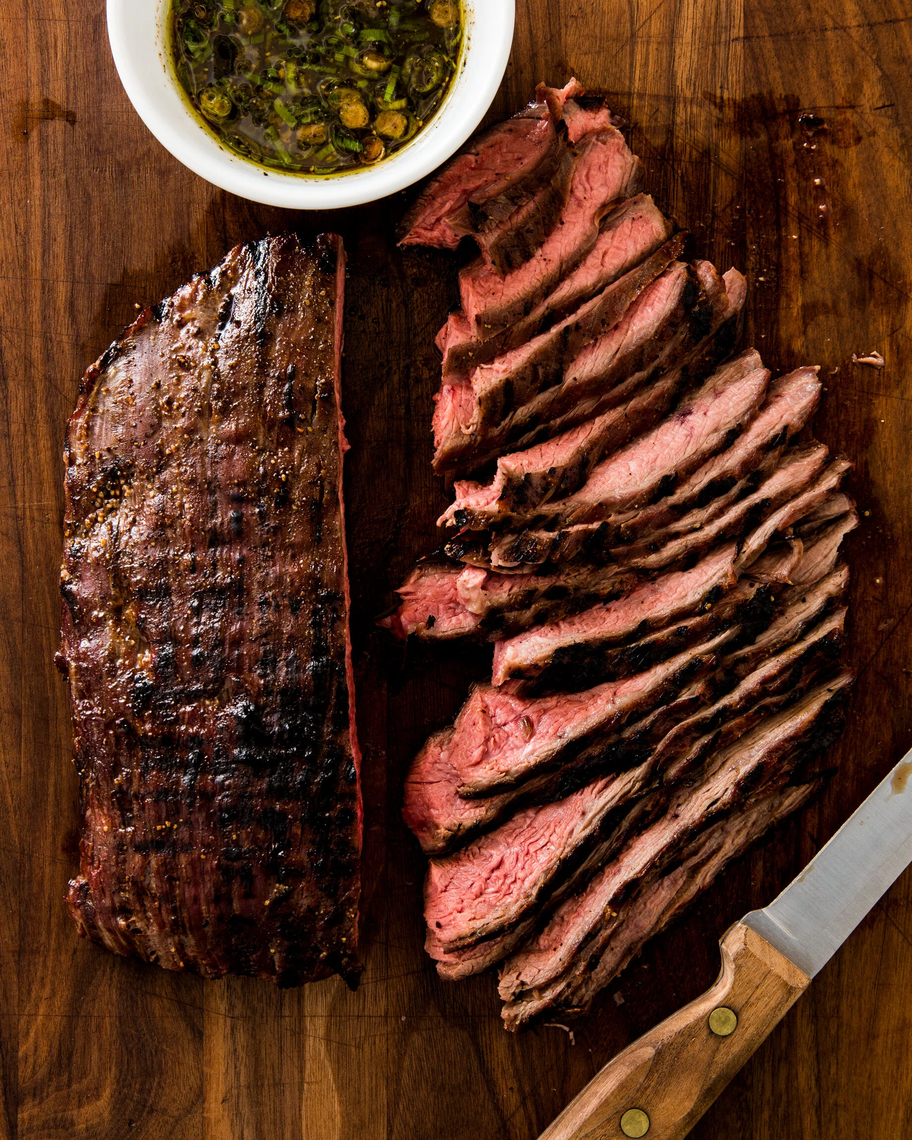Sliced grilled steak on a wooden cutting board with a side of chimichurri sauce in a bowl. Boston based food and hospitality photographer.