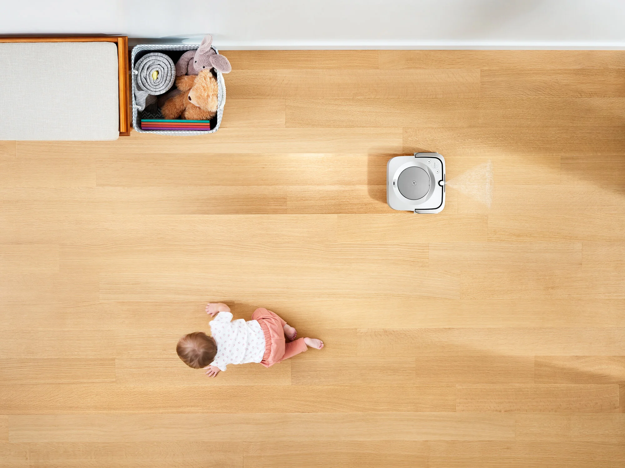 A baby crawling on a wooden floor near a white robotic vacuum cleaner, with a basket of plush toys and folded blankets in the corner. Photo created for an iRobot marketing campaign. 