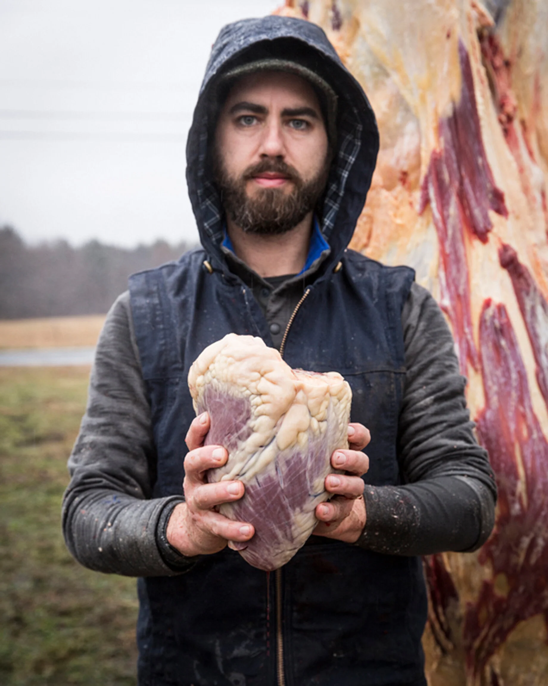 A bearded man in rain gear holding a large piece of raw meat outdoors with a foggy, open field in the background.