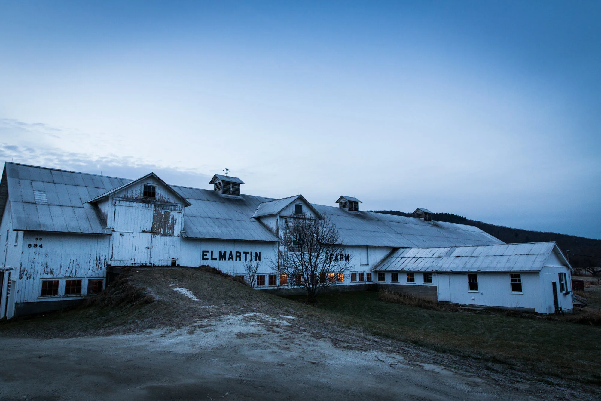 Old white farm building with the words 'Elmartin Farm' on it, situated on a small hill under a blue sky at dusk.