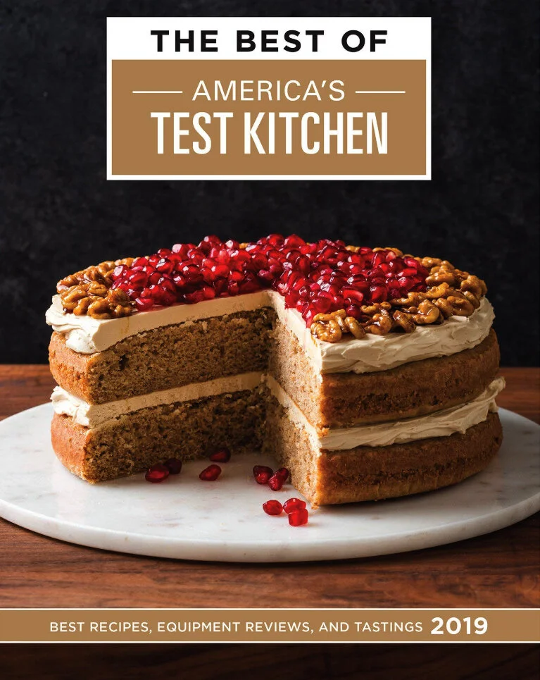 Magazine cover titled 'The Best of America's Test Kitchen 2019' featuring a sliced three-layer cake with white frosting, pomegranate seeds, and walnuts on top, set on a white marble cake stand against a dark background.