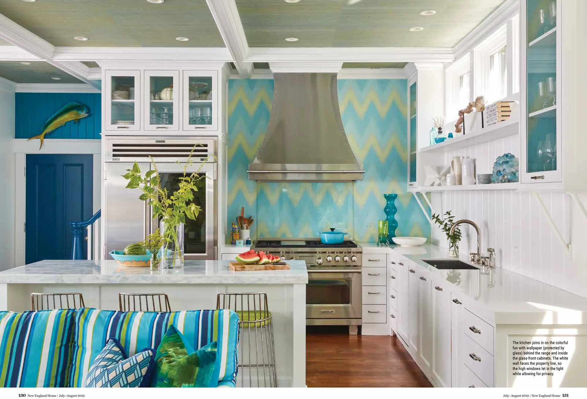 Colorful kitchen with white cabinets, stainless steel appliances, blue and green painted wall, and a kitchen island with sliced watermelon. Bright sunlight streams through windows, illuminating the space.