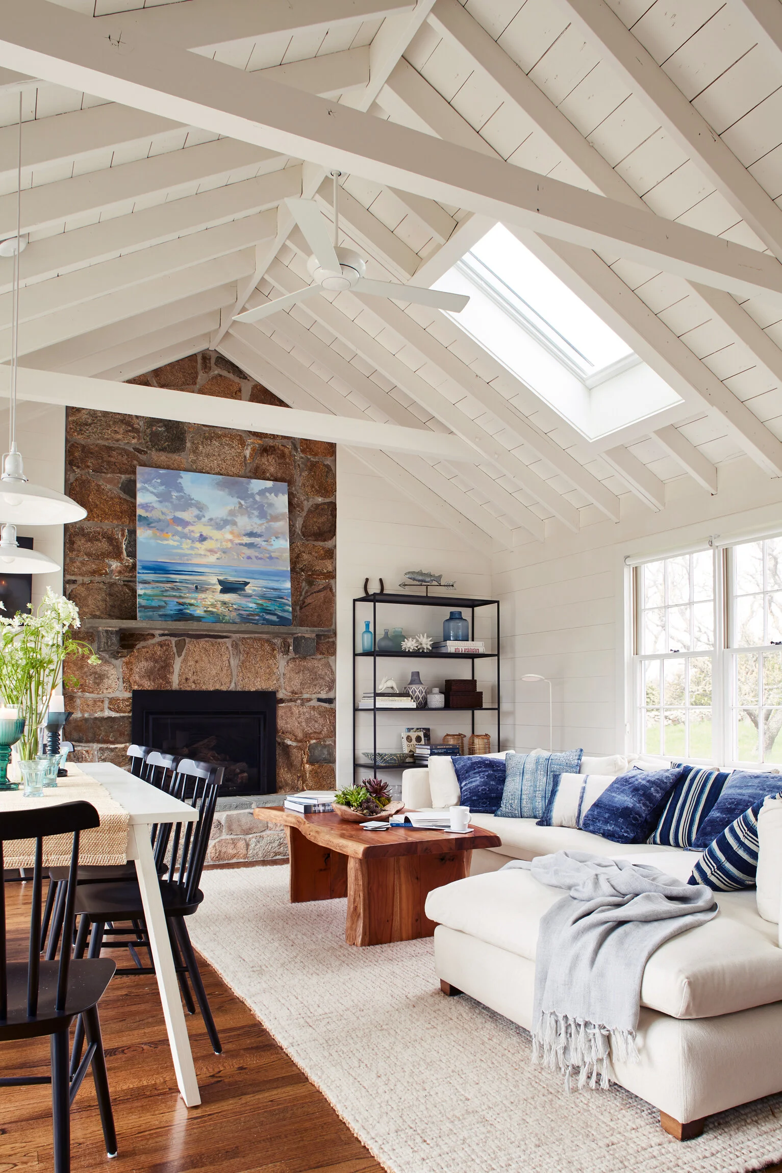 Living room with white vaulted ceiling, skylight, stone fireplace with painting above, white sofa with blue pillows, wooden coffee table, black bookshelf, and large windows.