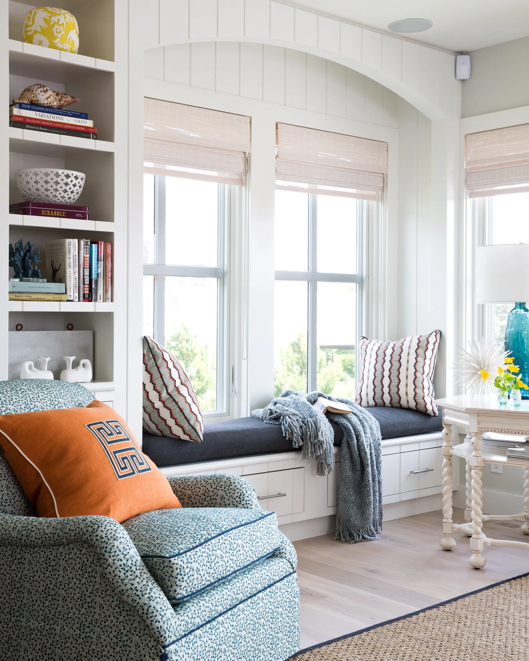 Cozy sunlit window nook with pillows and a blanket, adjacent to a patterned armchair and a white side table with a blue vase and flowers.