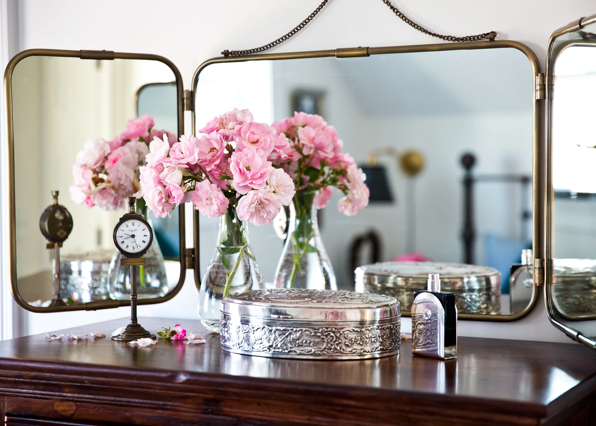 A wooden dresser with three silver ornaments, a glass vase with pink flowers, a small vintage clock, and a silver jewelry box, reflected in a triptych mirror on the dresser. An additional mirror is visible on the right side.