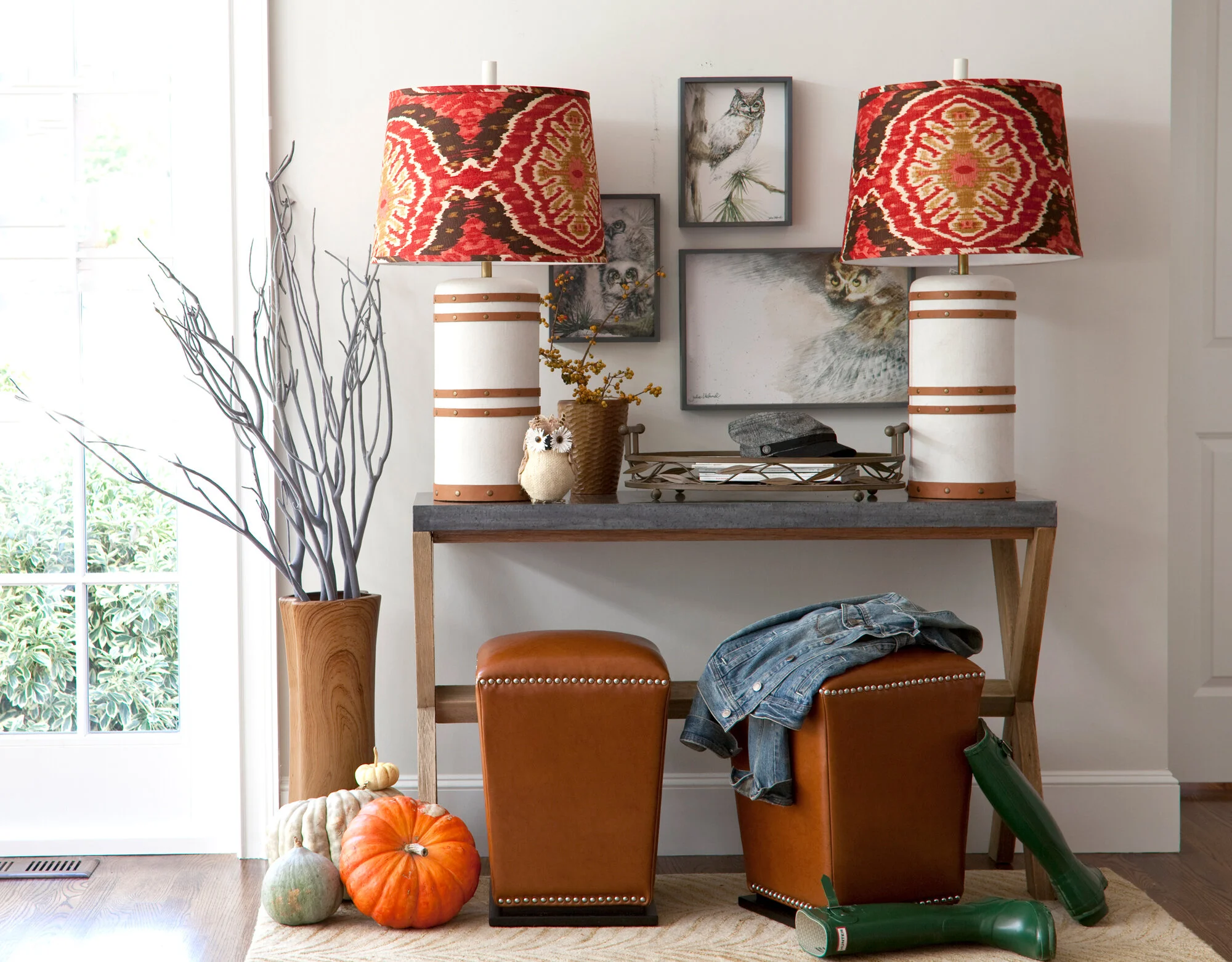 Living room decor with a wooden console table, two orange leather stools, a pair of textured table lamps with red patterned shades, wall art featuring cats and an owl, a vase with branches, pumpkins, and a pair of green rubber boots on the floor.