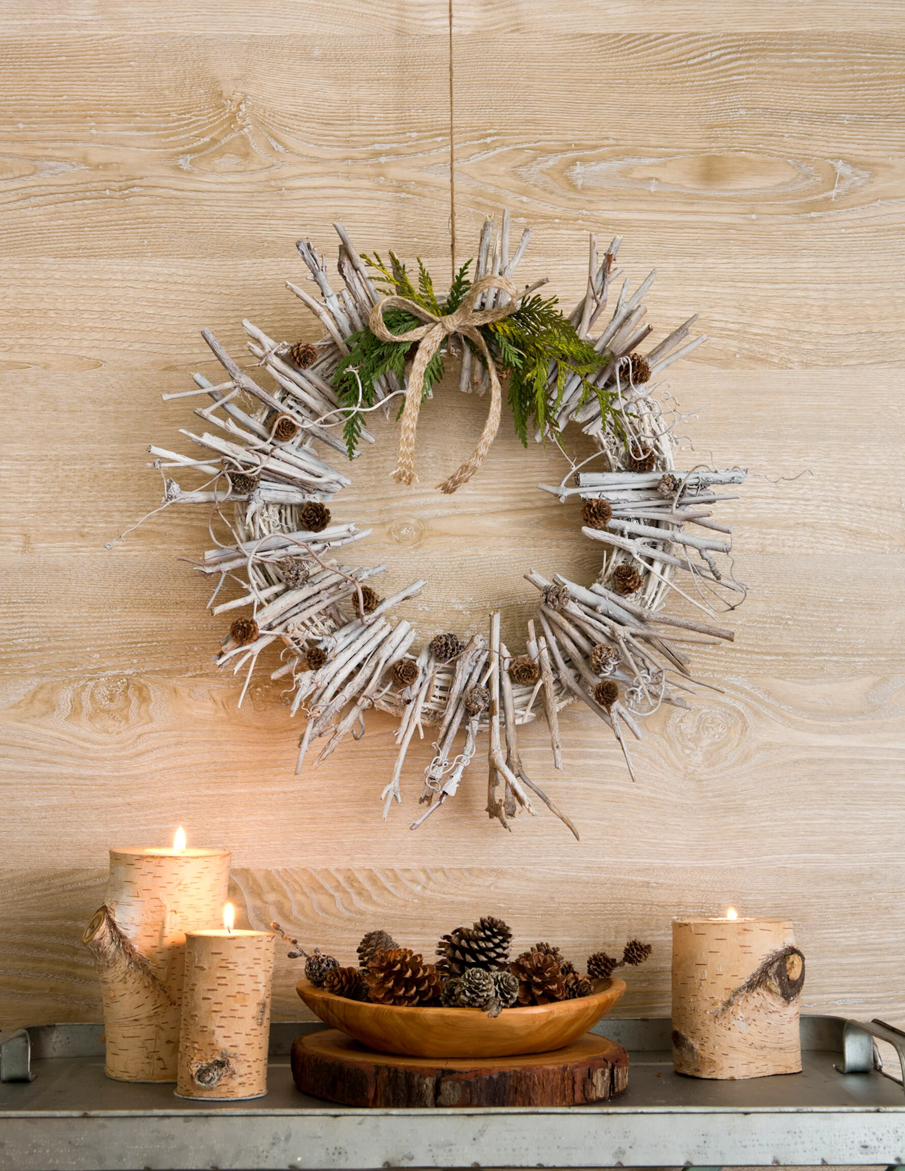 Rustic holiday decor featuring a white driftwood wreath with pinecone accents and a bow, flanked by three carved birch candle holders with lit candles, in front of a wooden wall with pinecones in a wooden bowl on a wooden slab.