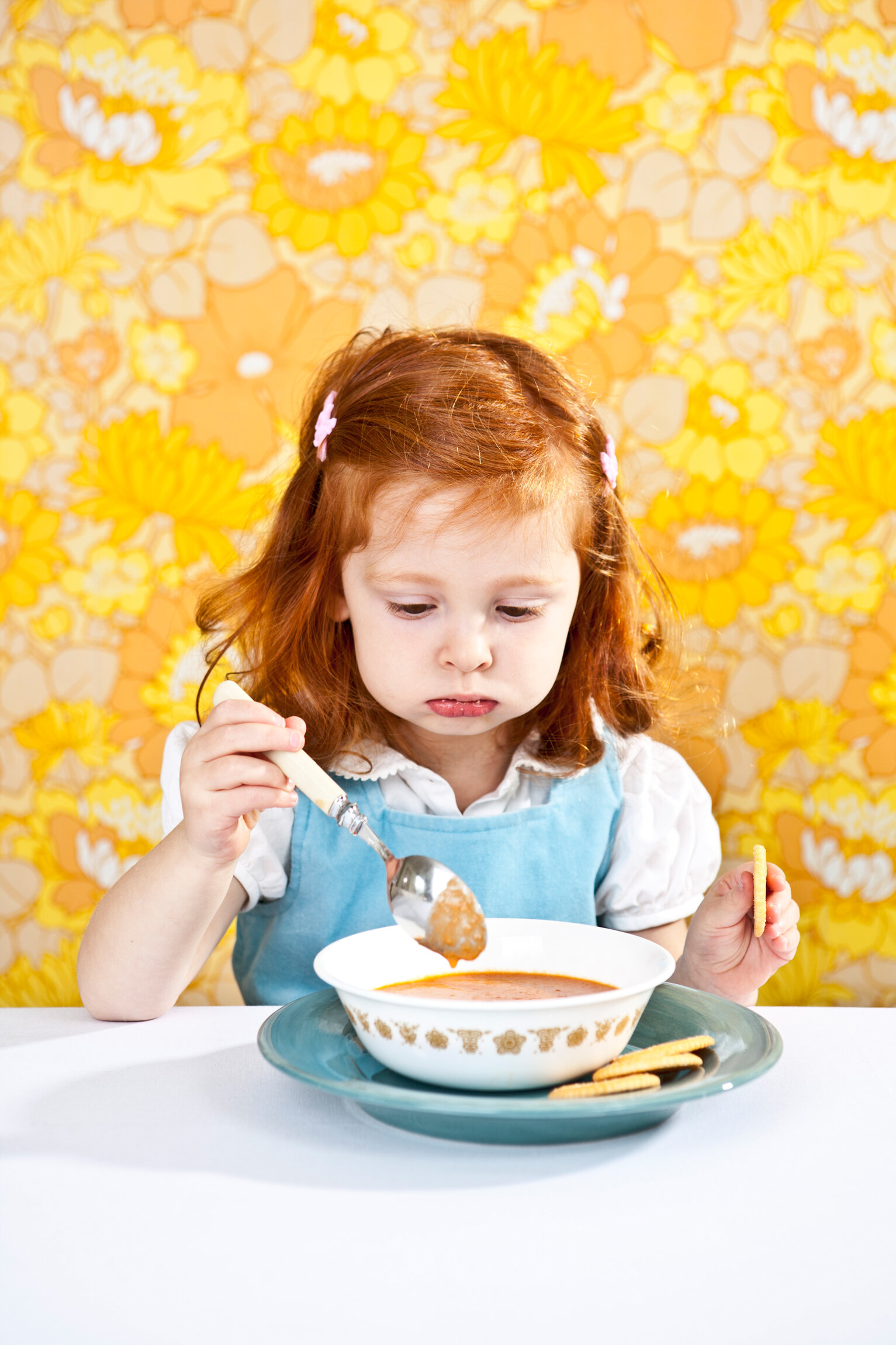 Young girl with red hair and pink hair clips sitting at a table, eating tomato soup with crackers against a yellow floral wallpaper background.