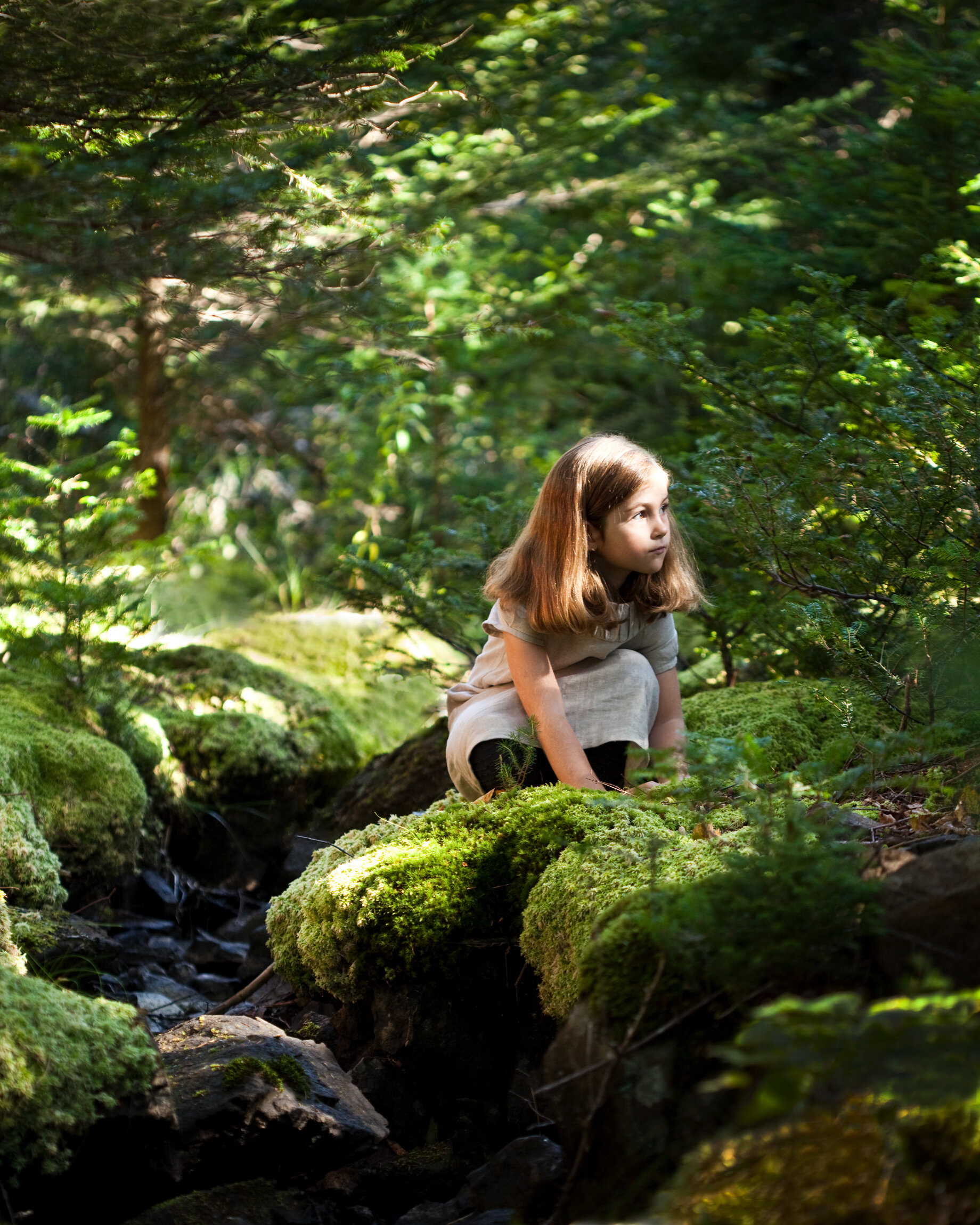 A young girl with brown hair wearing a beige dress squatting on moss-covered rocks in a lush green forest, looking intently to her right.