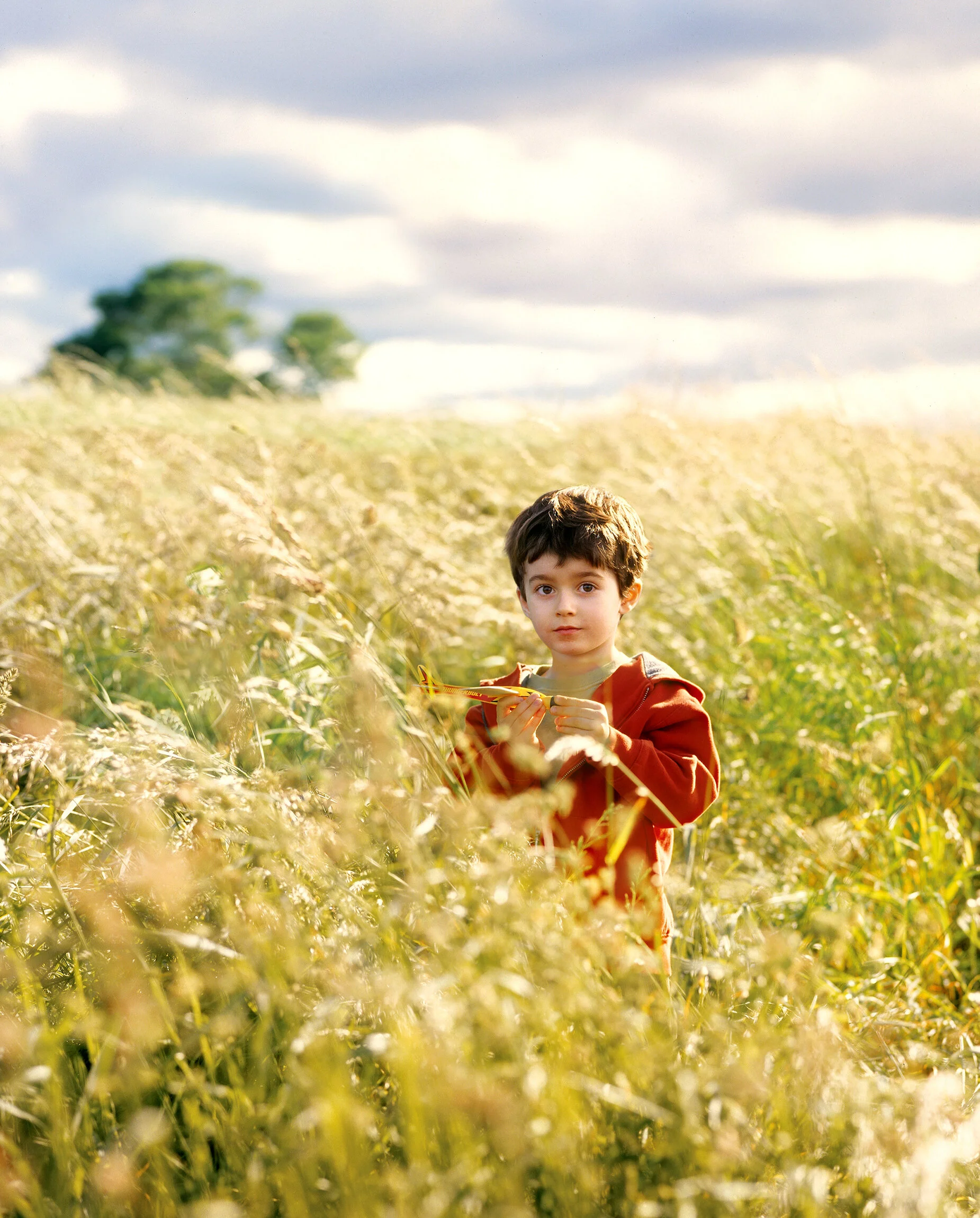 A young boy with dark hair standing in a grassy field holding a yellow toy airplane, wearing a red jacket, with a cloudy sky and trees in the background.