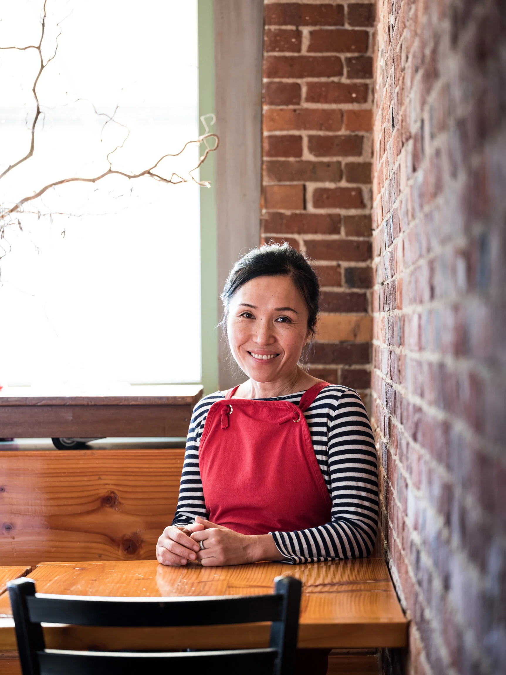 Woman with dark hair in a ponytail smiling, wearing a red apron over a striped shirt, sitting at a wooden table next to a brick wall in a restaurant or cafe.