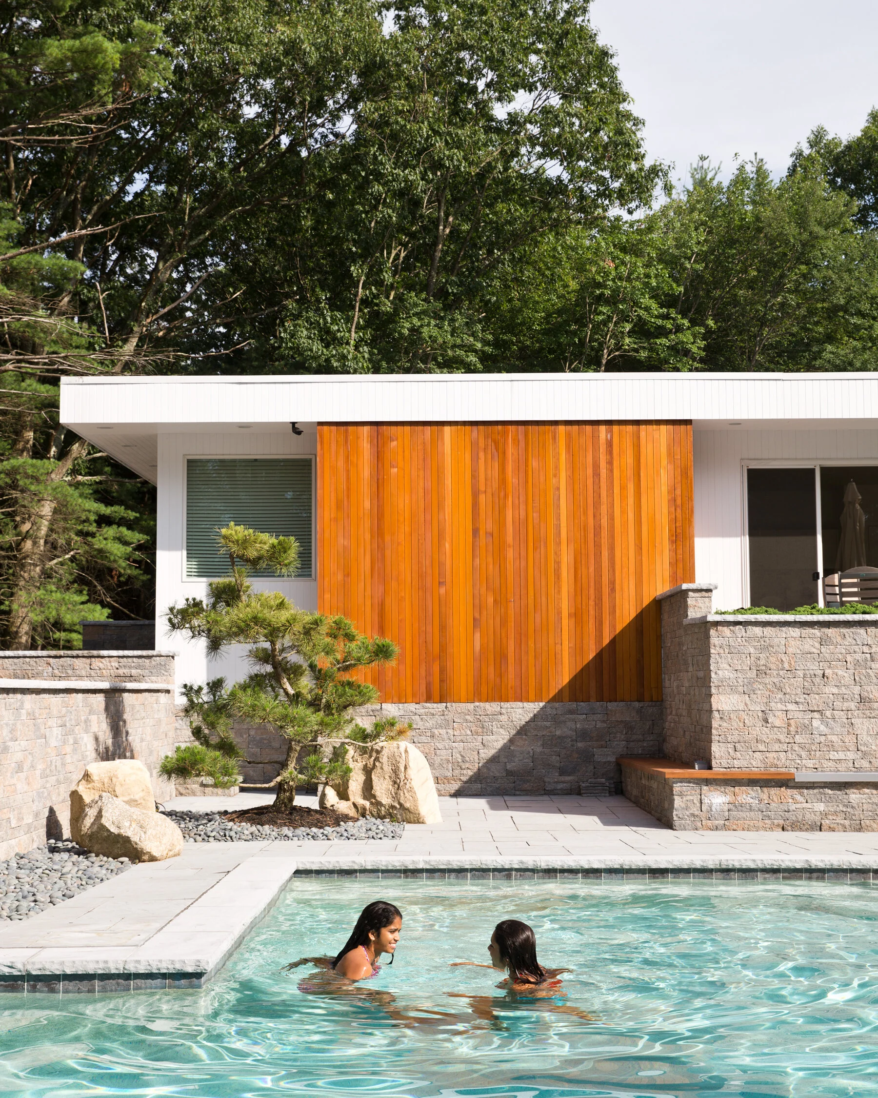 Two women swimming and talking in a backyard pool, with a modern house featuring a wooden accent wall and trees in the background.