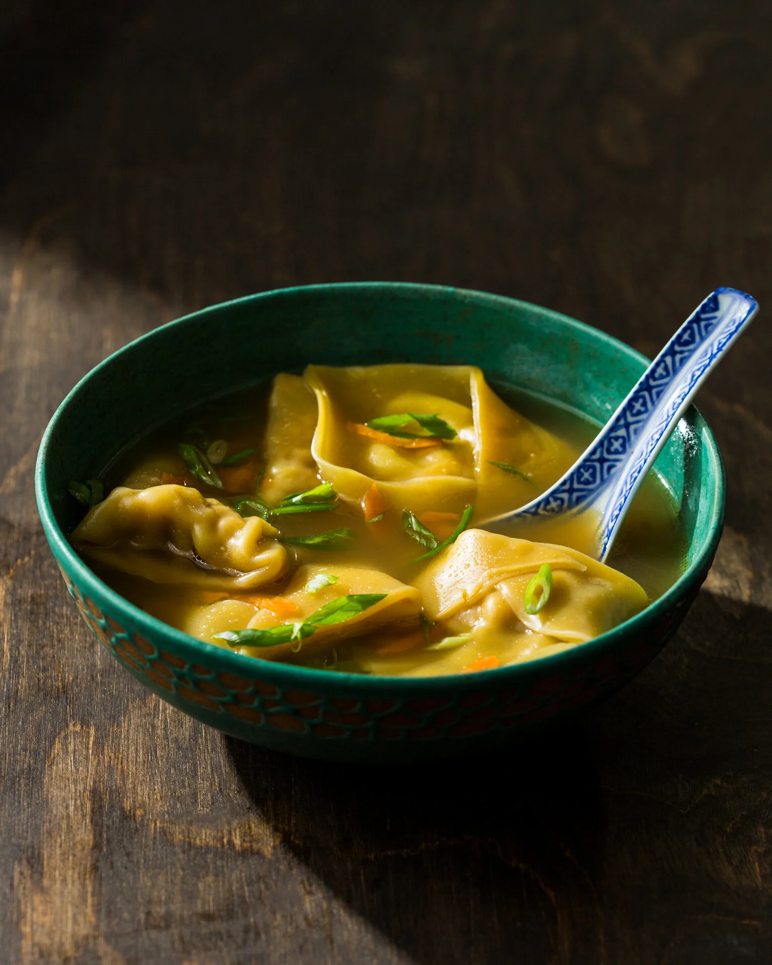 A bowl of Asian-style wonton soup with green onions and a patterned spoon on a dark wooden surface. Boston based food and hospitality photographer.