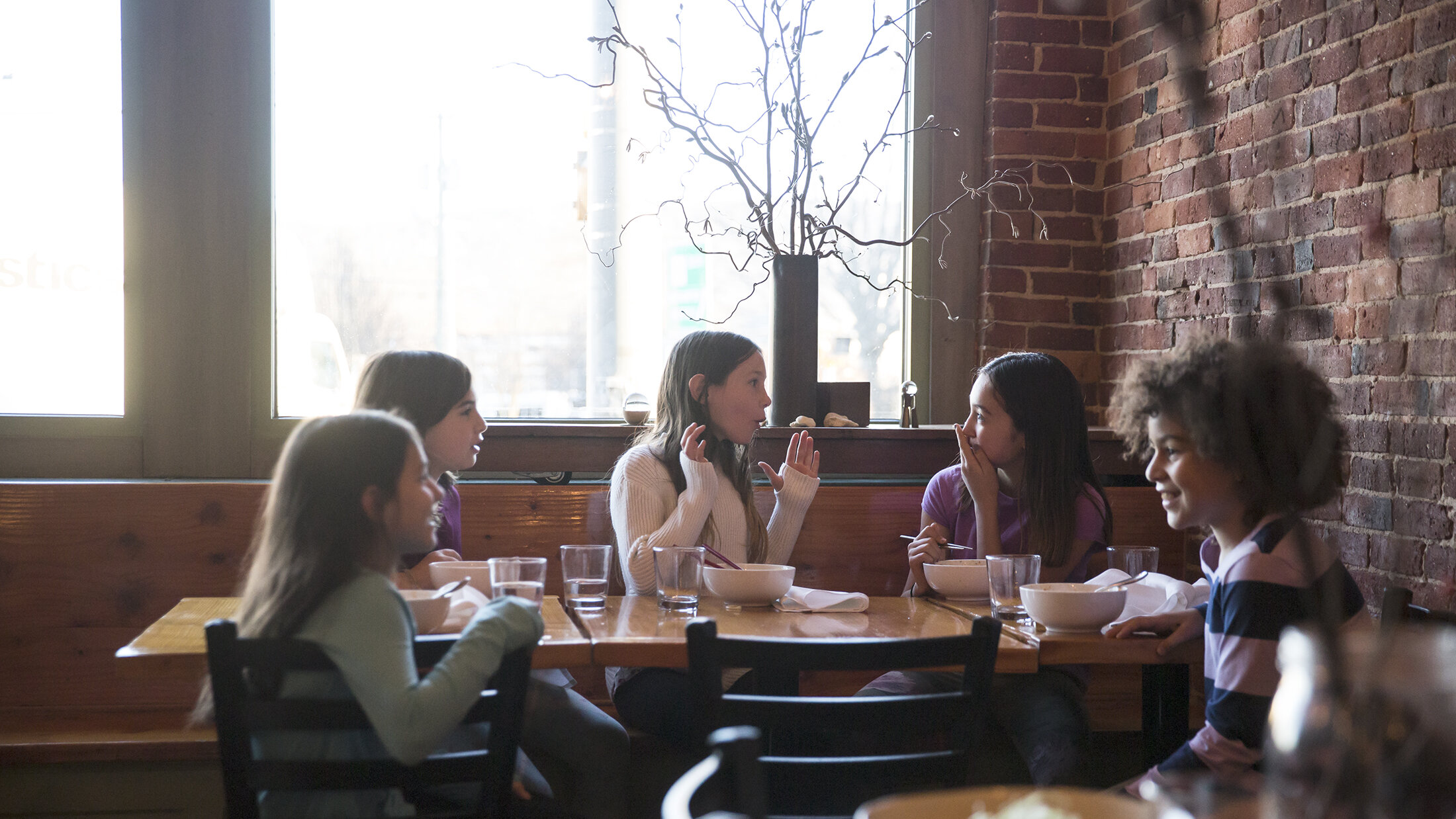 A group of five young girls sitting around a wooden table in a restaurant, talking and smiling, with large windows and a brick wall in the background.