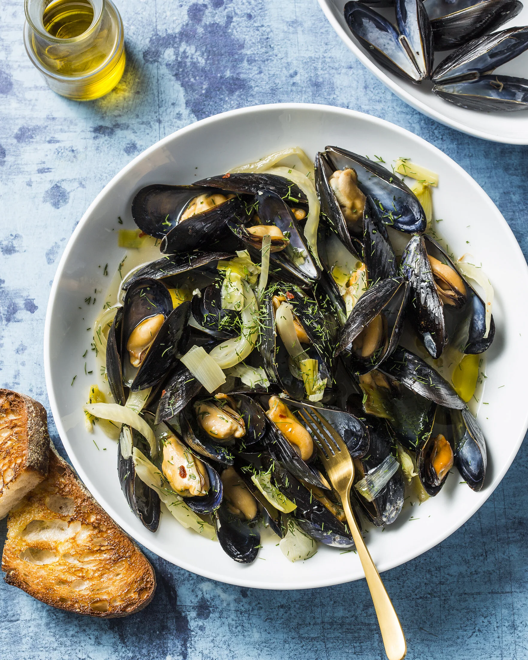 A white bowl filled with cooked mussels with herbs and vegetables on a blue textured surface, with a glass of white wine and a slice of toasted bread nearby. Boston based food and hospitality photographer.