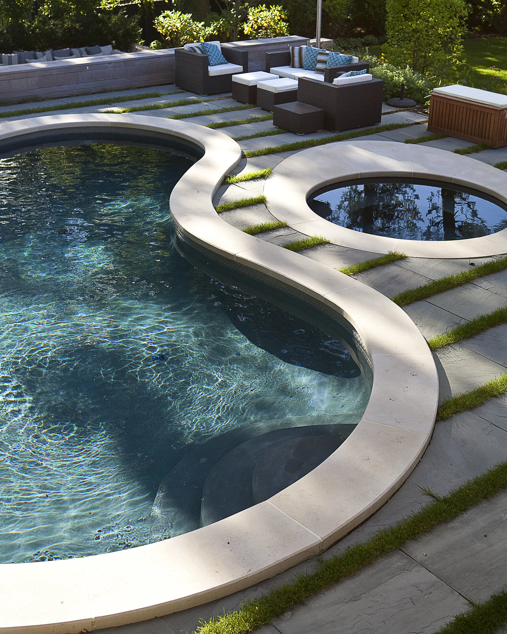 A backyard with a kidney-shaped swimming pool and a smaller round spa, surrounded by a stone patio and outdoor seating with cushioned chairs and pillows, and lush green trees in the background.