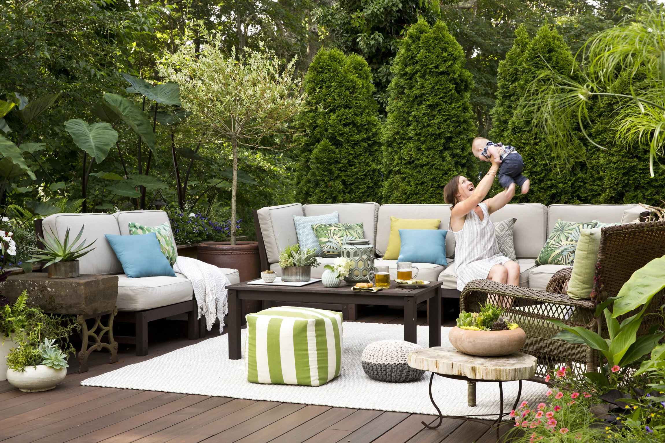 Woman in a white dress playing with a baby on a patio surrounded by lush green plants and outdoor furniture with colorful cushions.