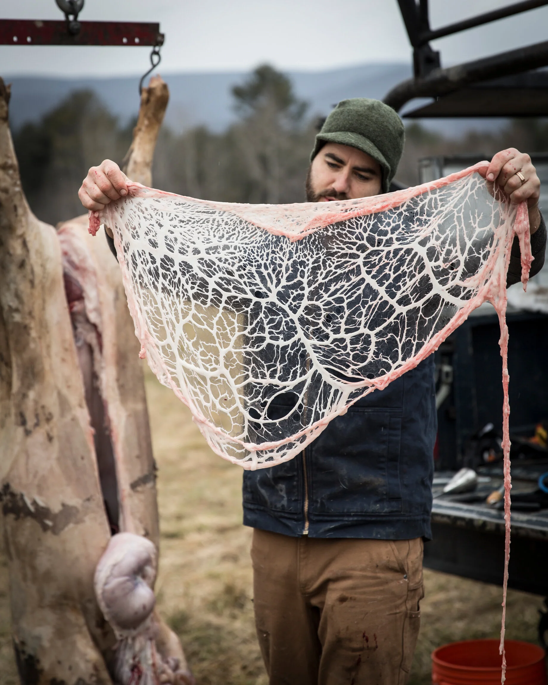 A man outdoors holds a large, delicate, lace-like sheet of animal fat or connective tissue. In the background, there is an animal carcass hanging, and the setting appears to be a rural or farm area.