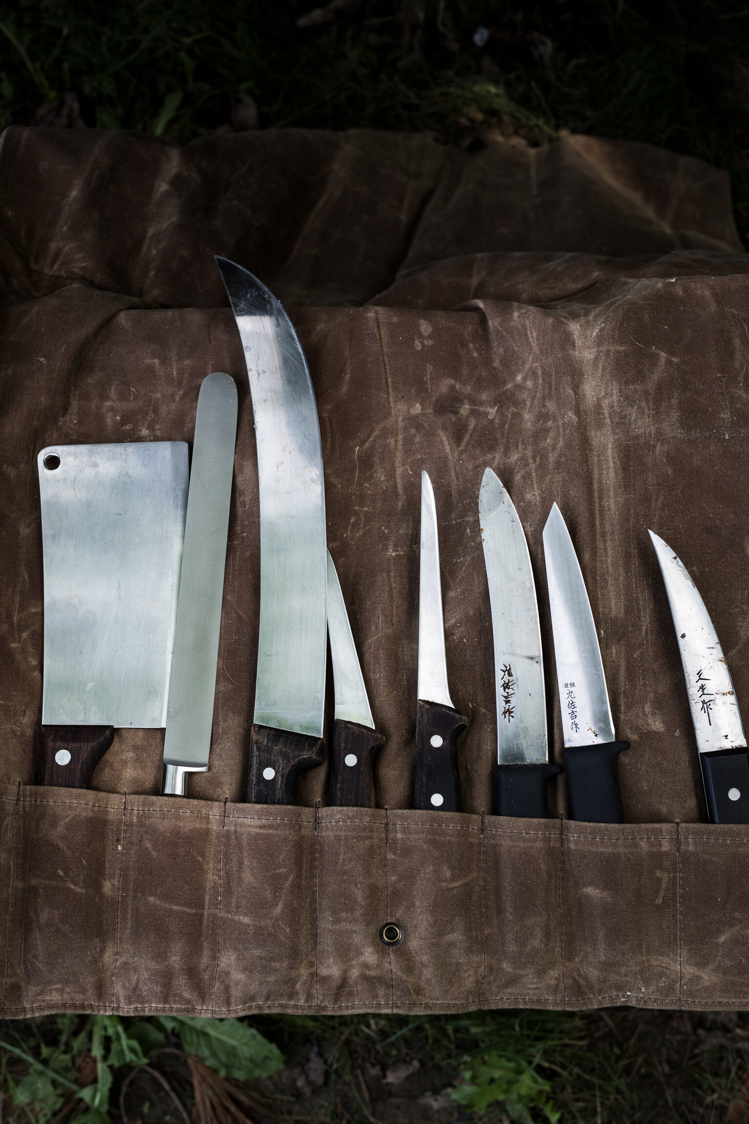 A set of eight kitchen knives with various sizes and shapes laid out on a brown fabric roll, outdoors on grass.