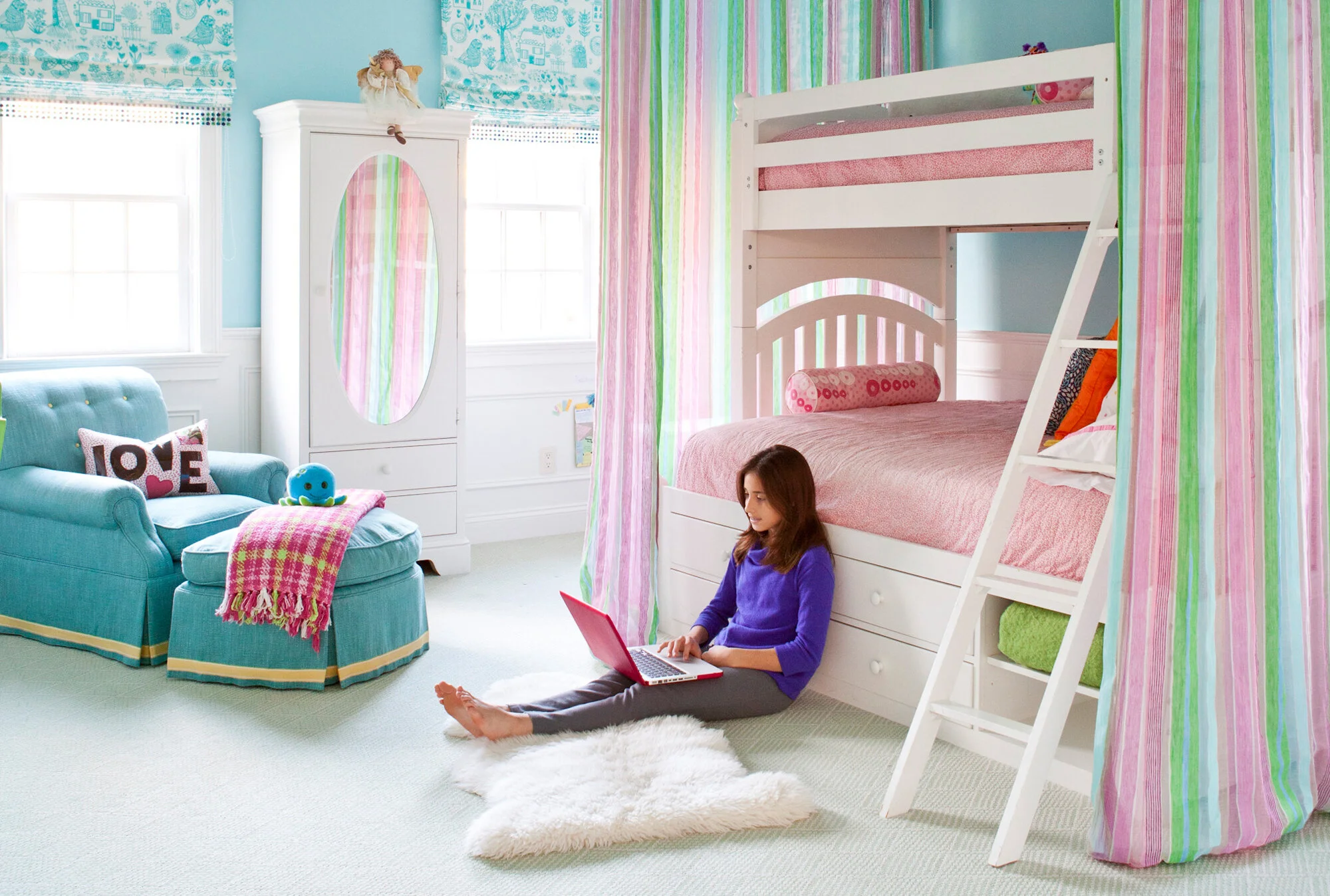 A girl sitting on a white fluffy rug with a pink laptop in a child's bedroom, which has pastel-colored decor, a bunk bed with pink bedding, a white wardrobe with an oval mirror, and a teal armchair with a pink and white pillow that says "LOVE" and a 
