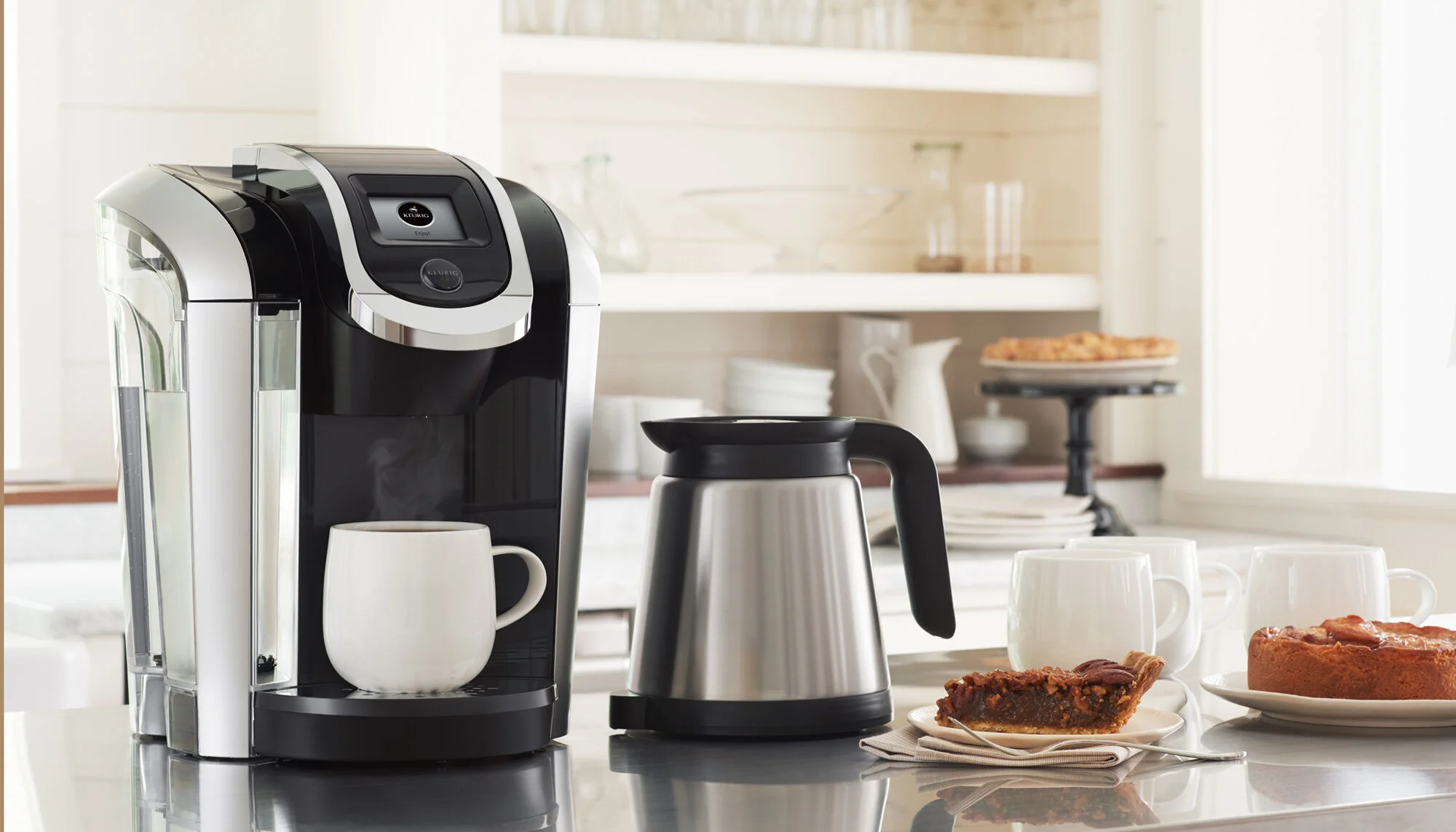 A kitchen countertop with a Keurig coffee maker brewing coffee into a mug, electric kettle, cups, and plates with pie and cake. Boston product photographer.