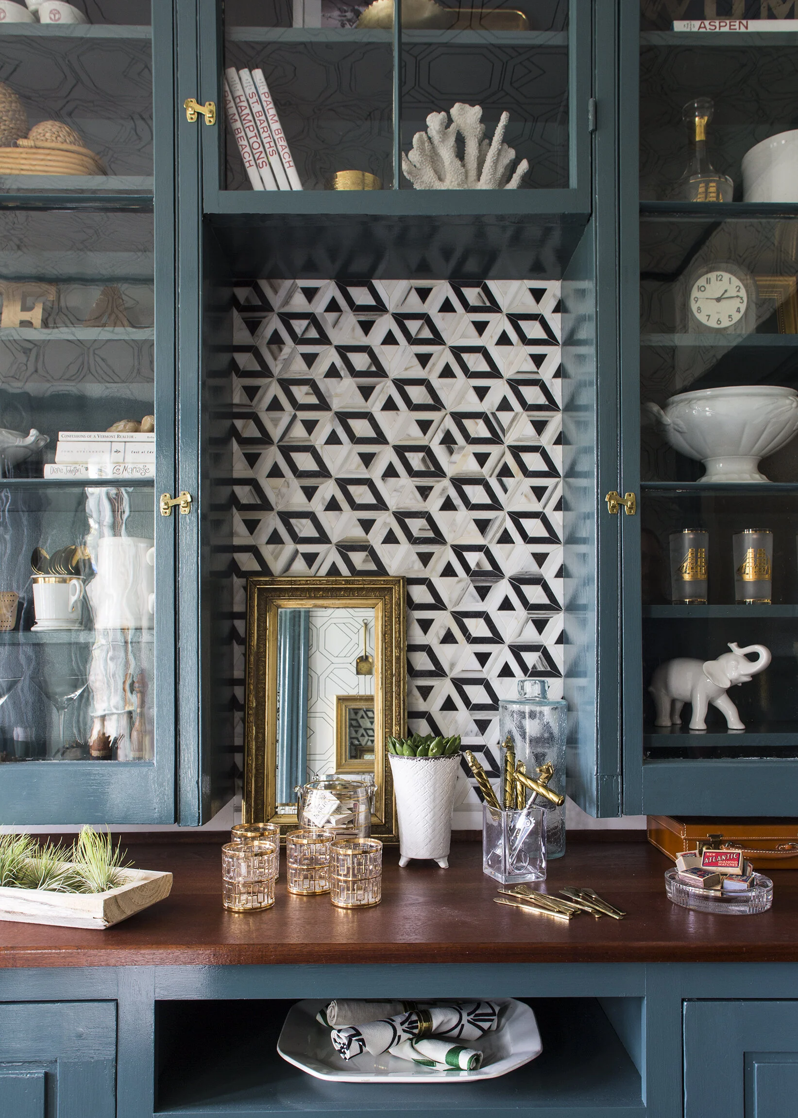 A kitchen cabinet with blue doors, decorative glassware, white and black geometric patterned backsplash, a small mirror with a gold frame, a white plant pot with a green succulent, gold utensils, and miscellaneous small items.