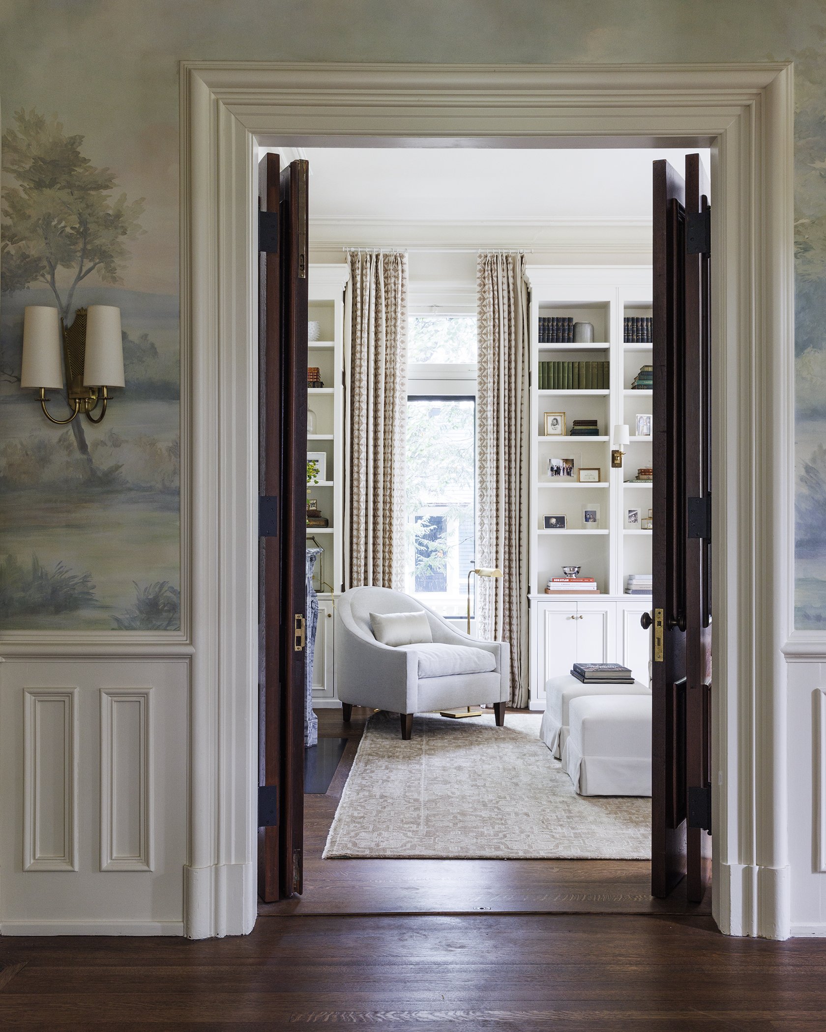 A view through open dark wood double doors into a bright living room with white built-in bookshelves, large window with striped curtains, a white armchair with a small pillow, a white ottoman, and a beige area rug.