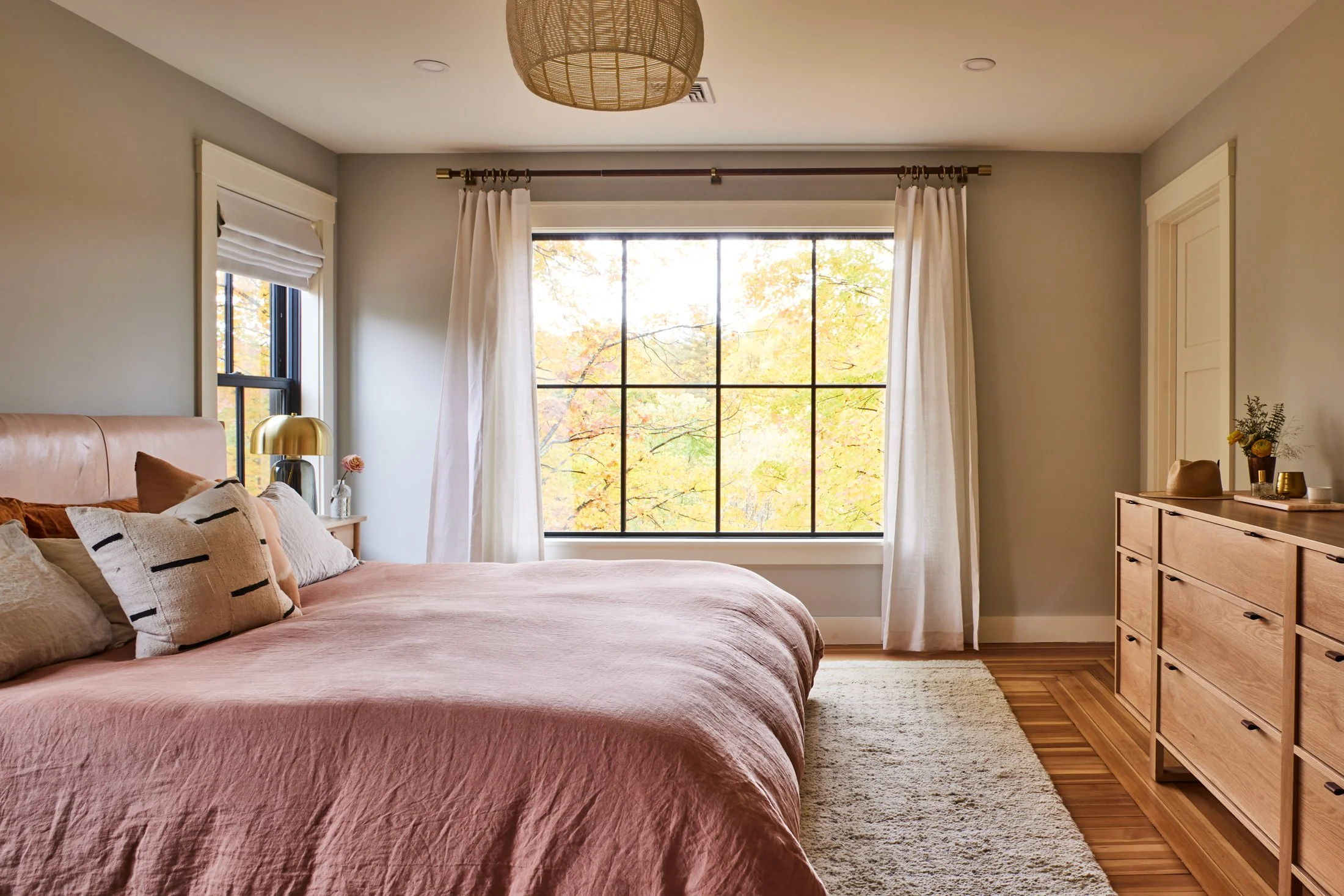 A cozy bedroom with a large window showing autumn trees, a pink bed with multiple pillows, a wooden dresser, and a beige rug on hardwood floor.