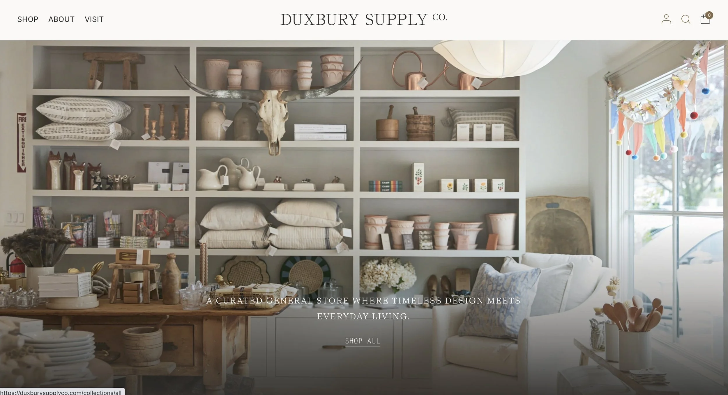 Interior view of a curated general store with shelves displaying various home decor items, ceramics, books, and textiles. Natural light filters in through a window decorated with colorful bunting.  Screenshot of Duxbury Supply Co's web site