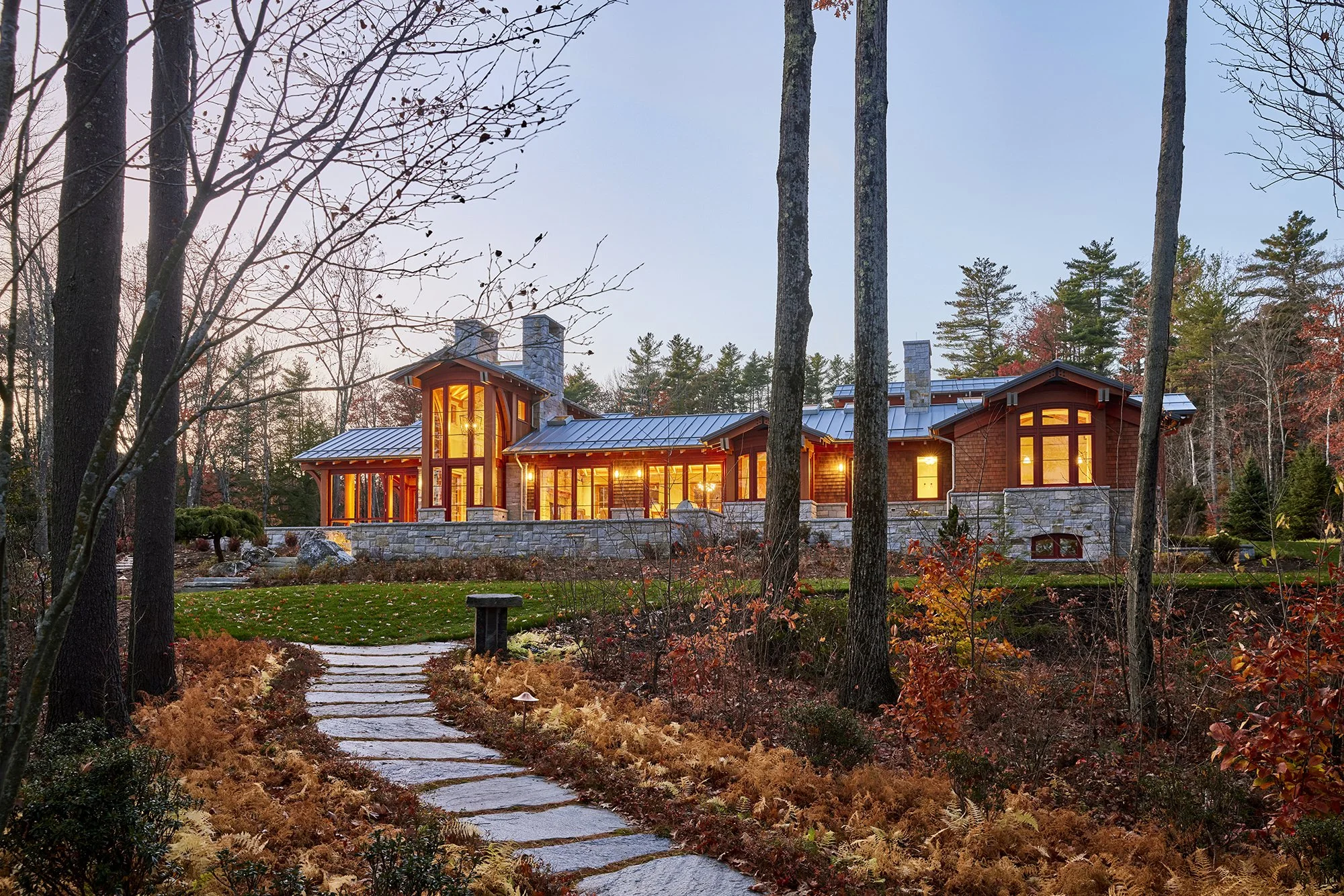 A modern house with large windows and stone accents, illuminated from within, situated in a wooded landscape with trees and autumn foliage during sunset. Professional architectural photograph of a custom home in Northern New England. 