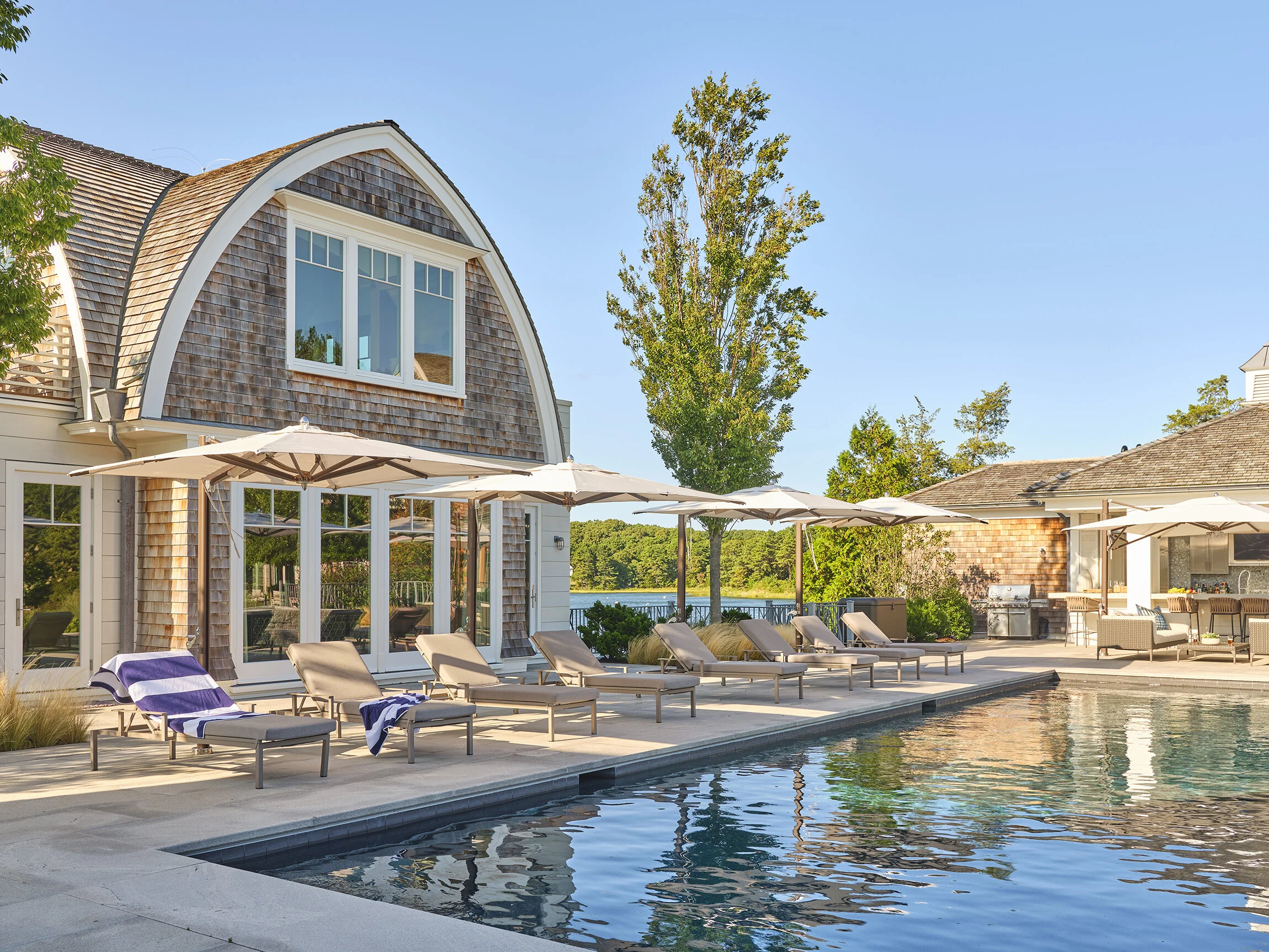 Poolside area with lounge chairs, umbrellas, and a house with large windows and wooden shingles in a backyard setting.