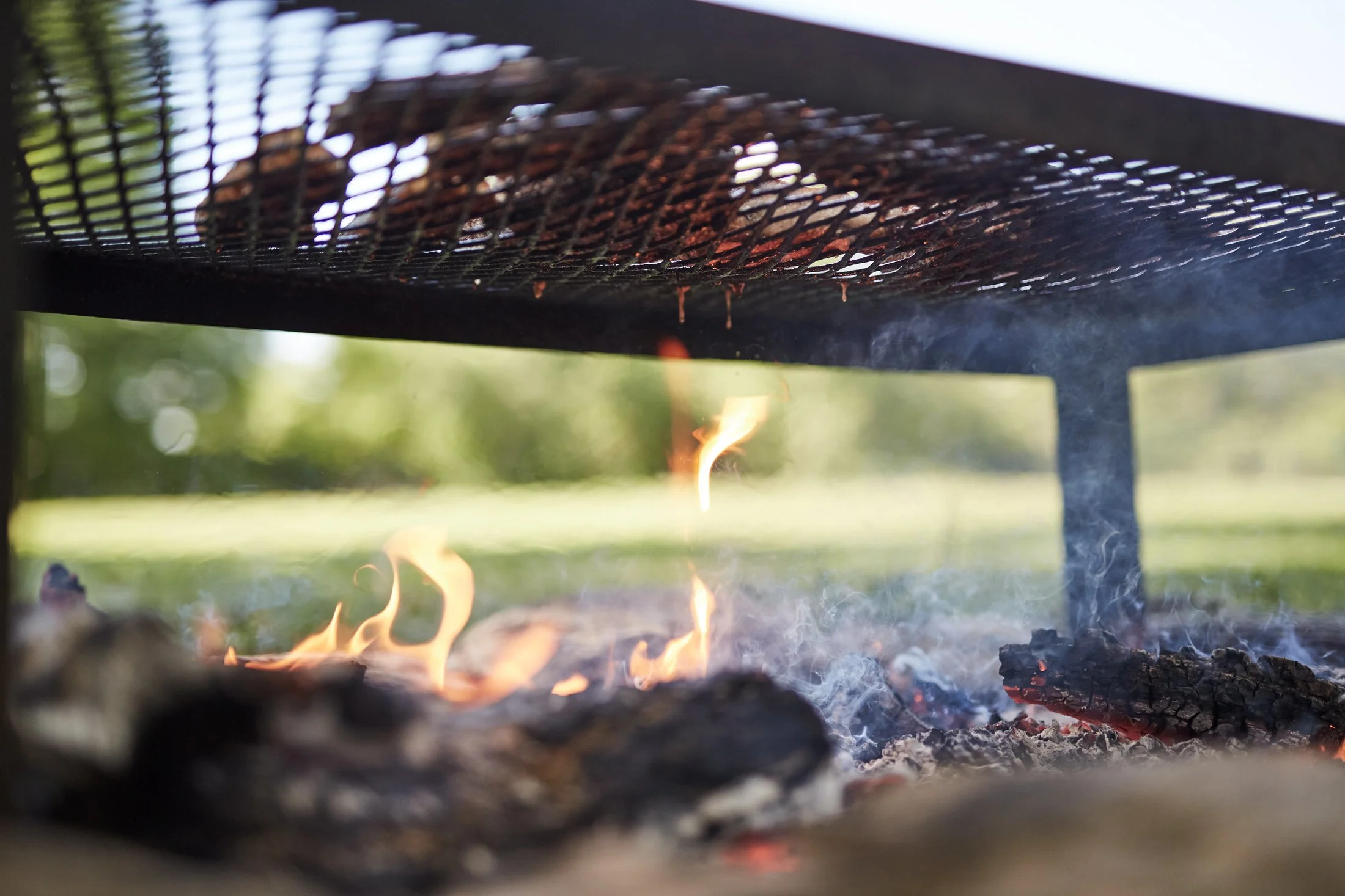 Close-up of a barbecue grill with burning wood underneath, with visible flames and smoke, set outdoors with a blurred green background.