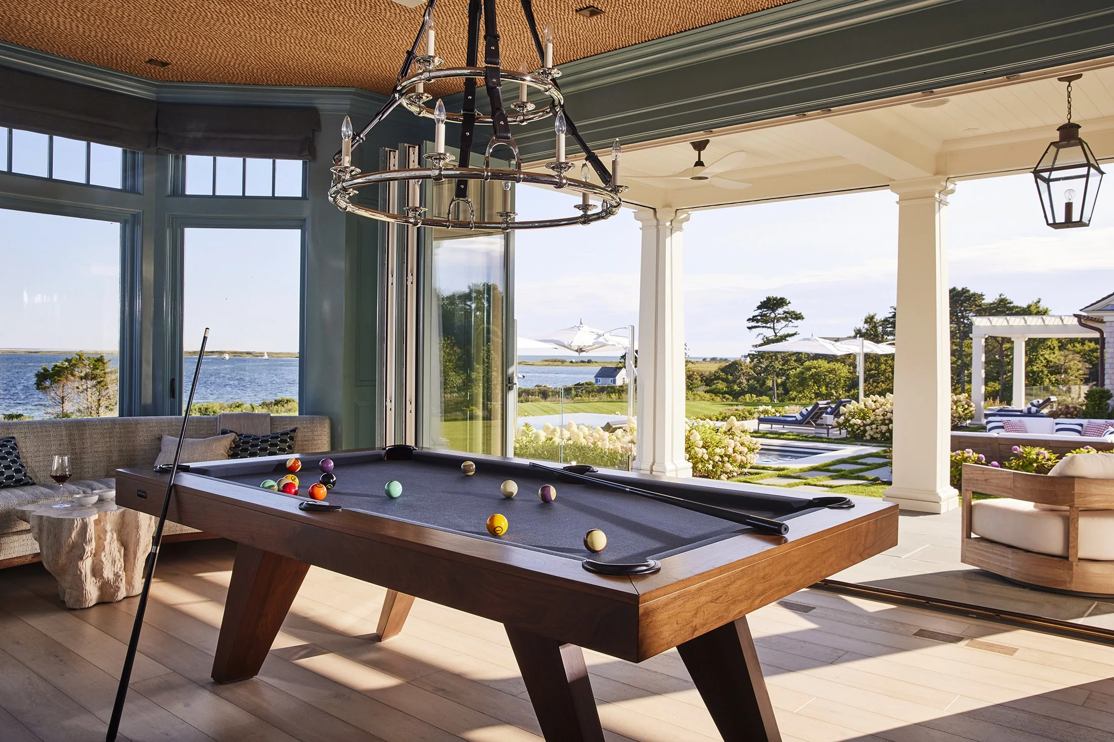 Indoor pool room with a billiards table, large windows overlooking a pool and outdoor landscape, open sliding doors, and outdoor chairs and umbrellas. Architectural photograph of a home on Cape Cod