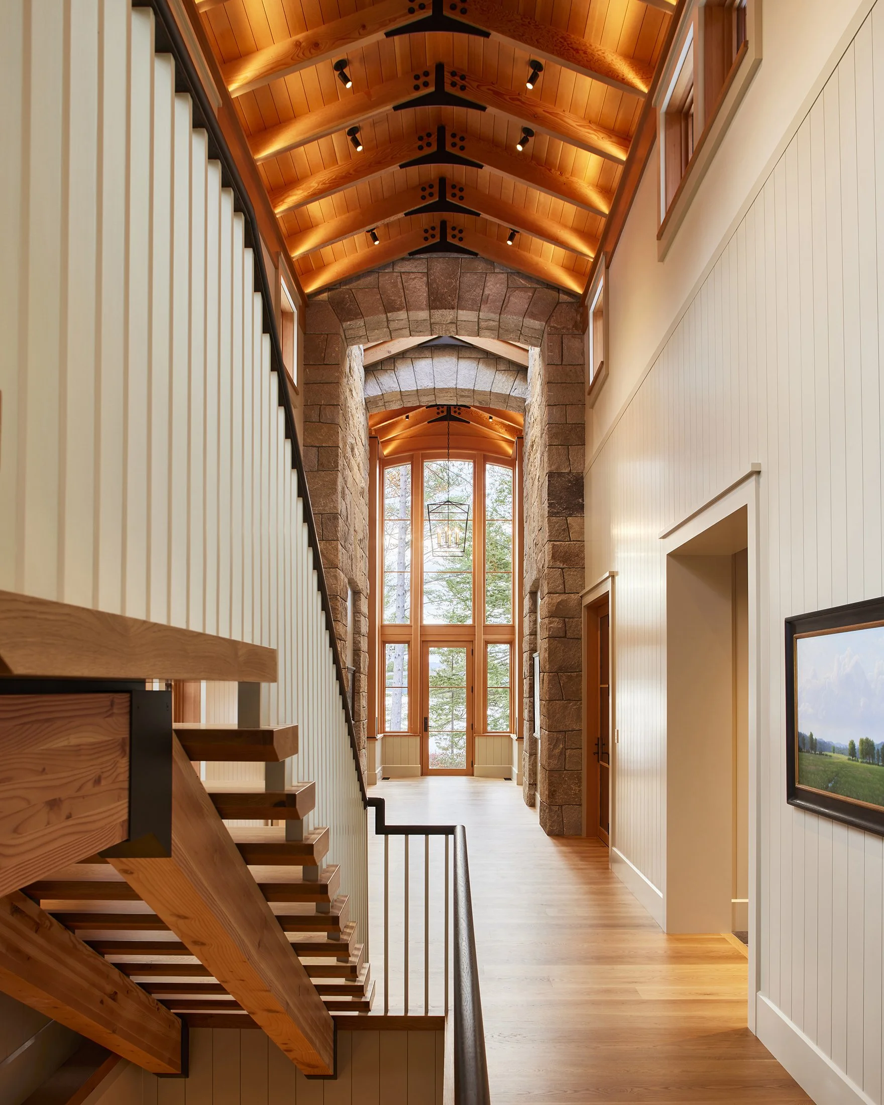Bright hallway with a high, arched wooden ceiling, a stone archway framing large glass doors at the end, and a wooden staircase on the left side leading upward. Light streams through the glass, illuminating the space. Professional architectural photo