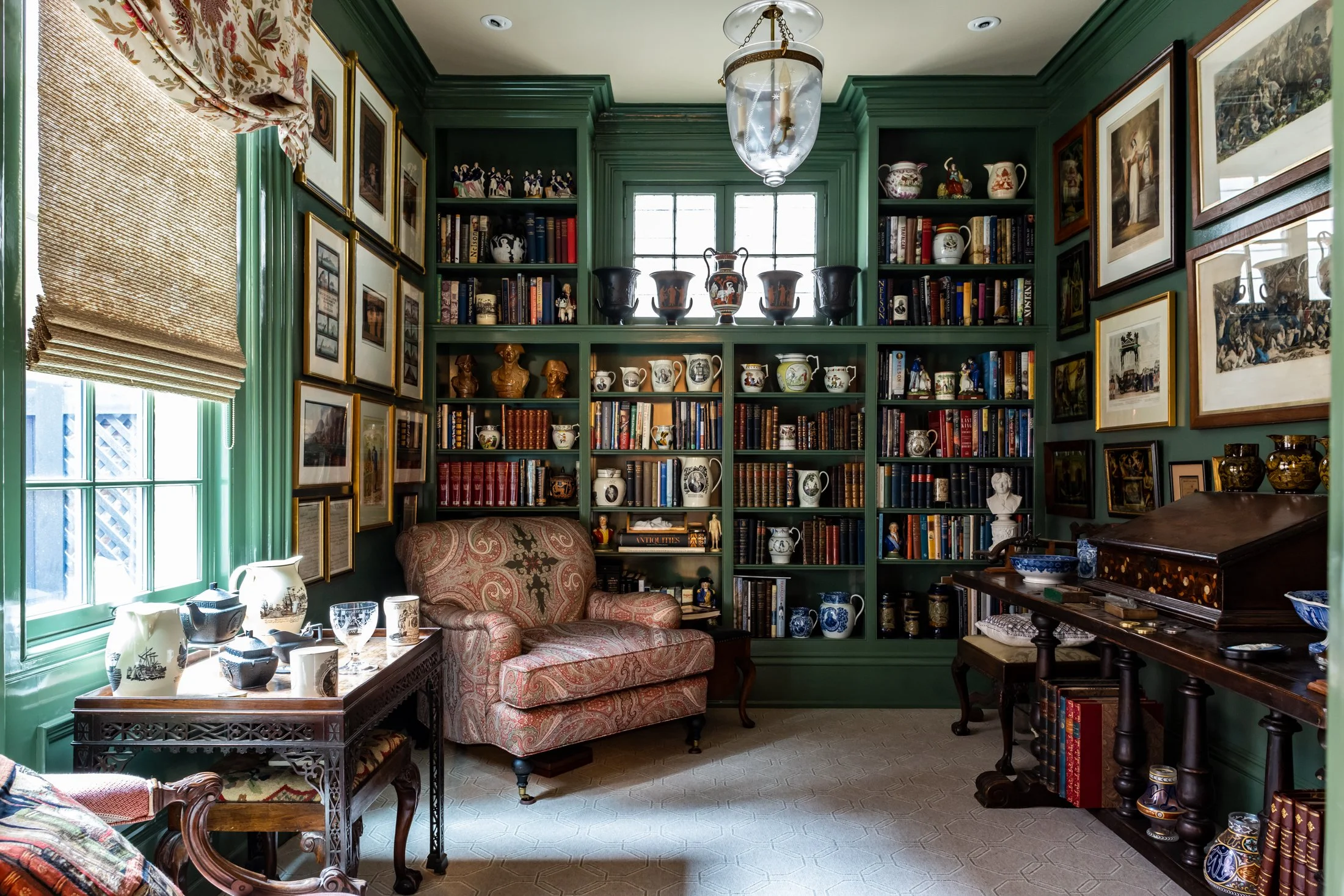 A cozy library room with green walls, filled with books, decorative ceramics, paintings, and bust sculptures, featuring a cushioned armchair, a window with a Roman shade, and a wooden side table with ceramic vases.