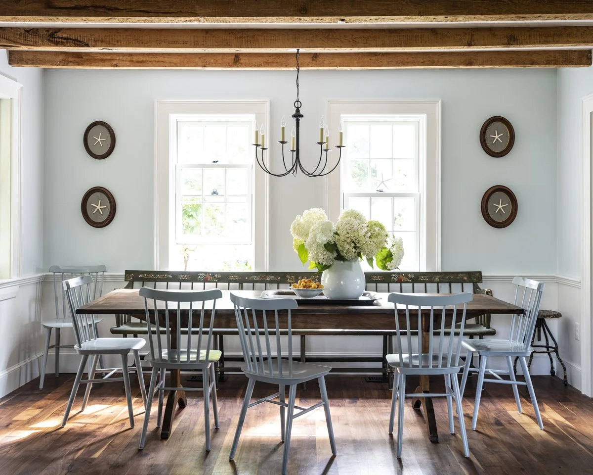 A dining room with a wooden table surrounded by white chairs, a large white flower vase in the center, two windows, a chandelier hanging from a wooden ceiling, and framed starfish decorations on the wall.