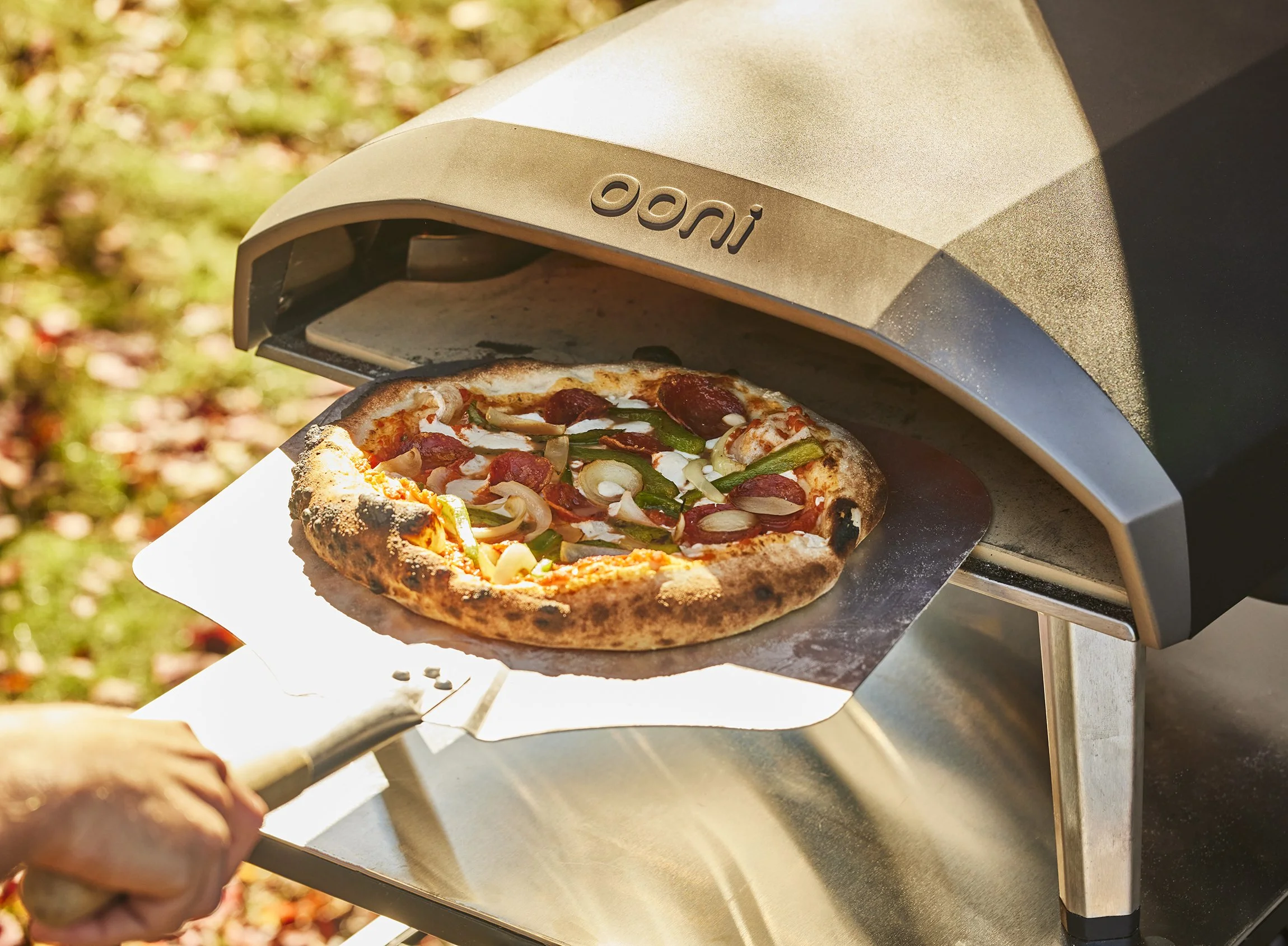 Pizza being cooked in a pizza oven outdoors, with toppings including pepperoni, onions, and green peppers.