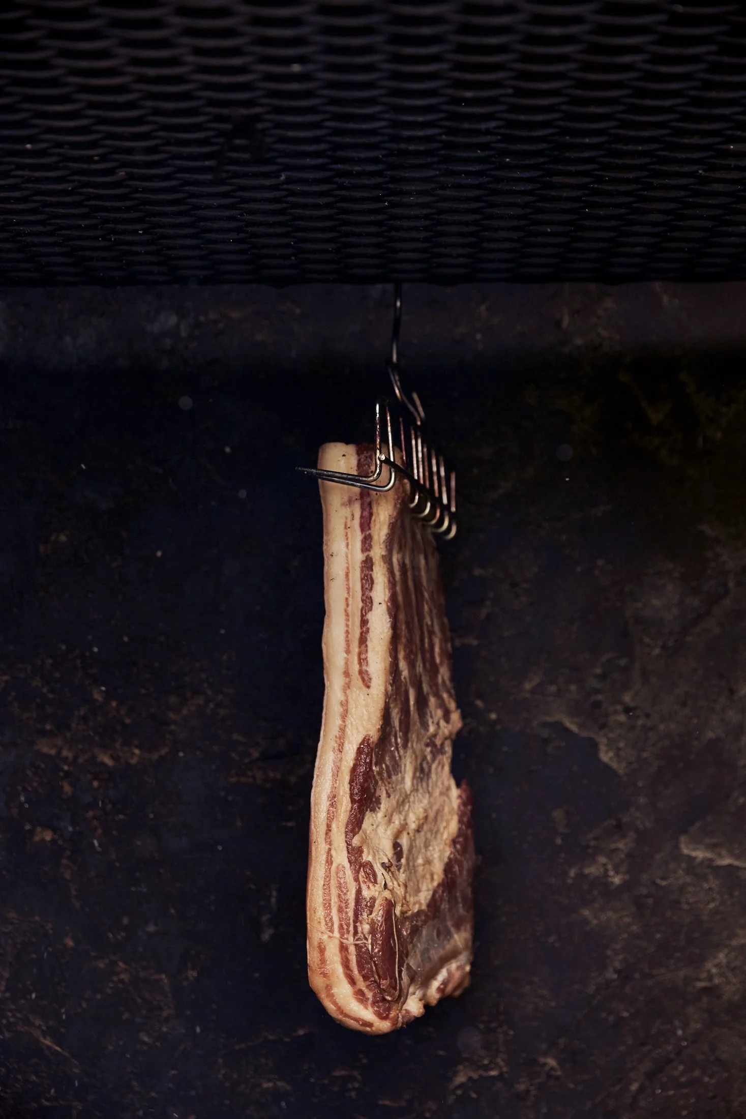 A strip of raw bacon hanging from a metal hook in a smoker or dry curing environment.