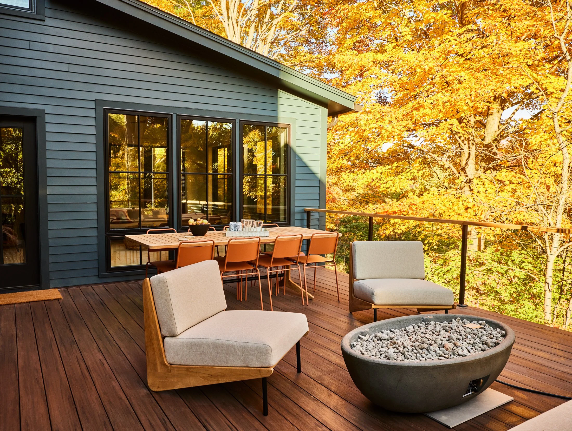 A spacious outdoor wooden deck with modern furniture, including two beige cushioned chairs and a fire pit filled with rocks. There is a rectangular dining table with six orange chairs. The deck is adjacent to a house with gray siding and large black-