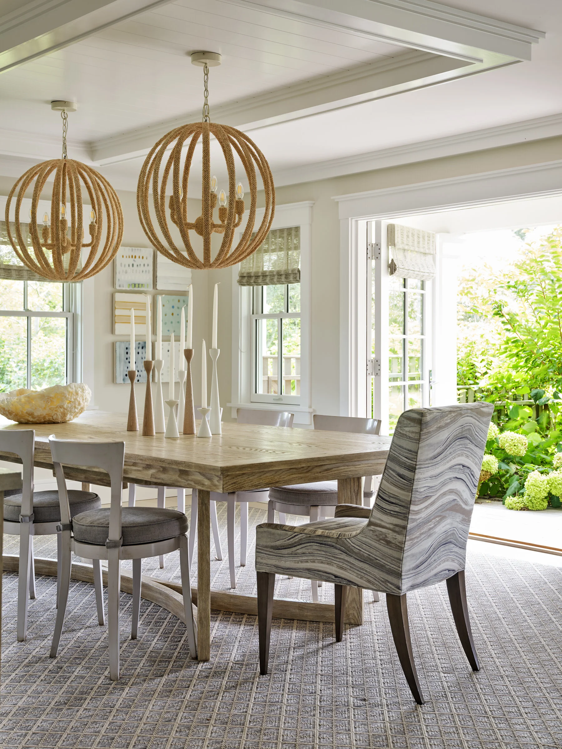 Bright dining room with a wooden table, upholstered and wooden chairs, decorative candle holders, modern woven pendant lights, large windows with Roman shades, and greenery outside.
