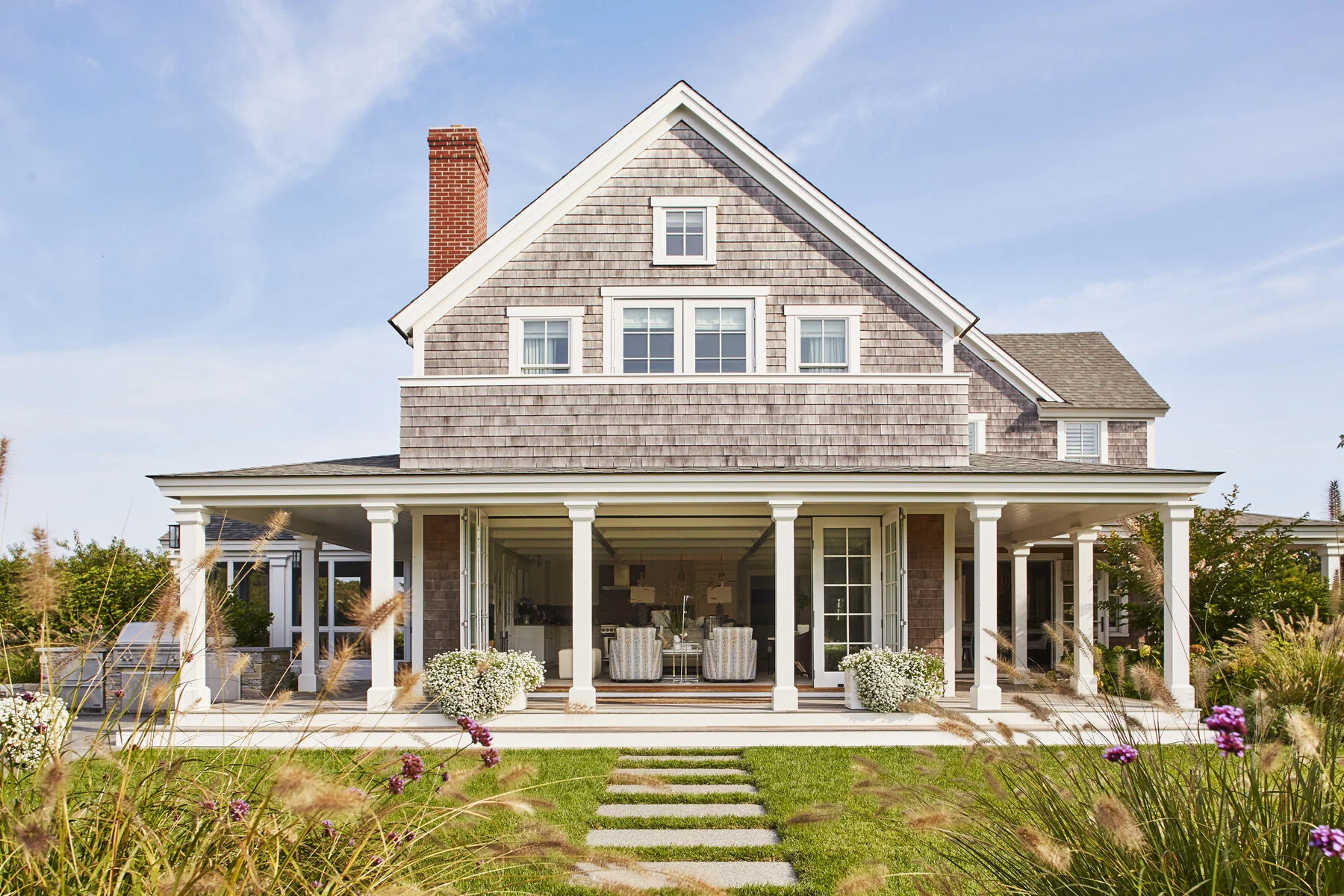 A two-story house with a wraparound porch, light brown shingles, white trim, and a brick chimney, surrounded by landscaped garden with colorful flowers and greenery.