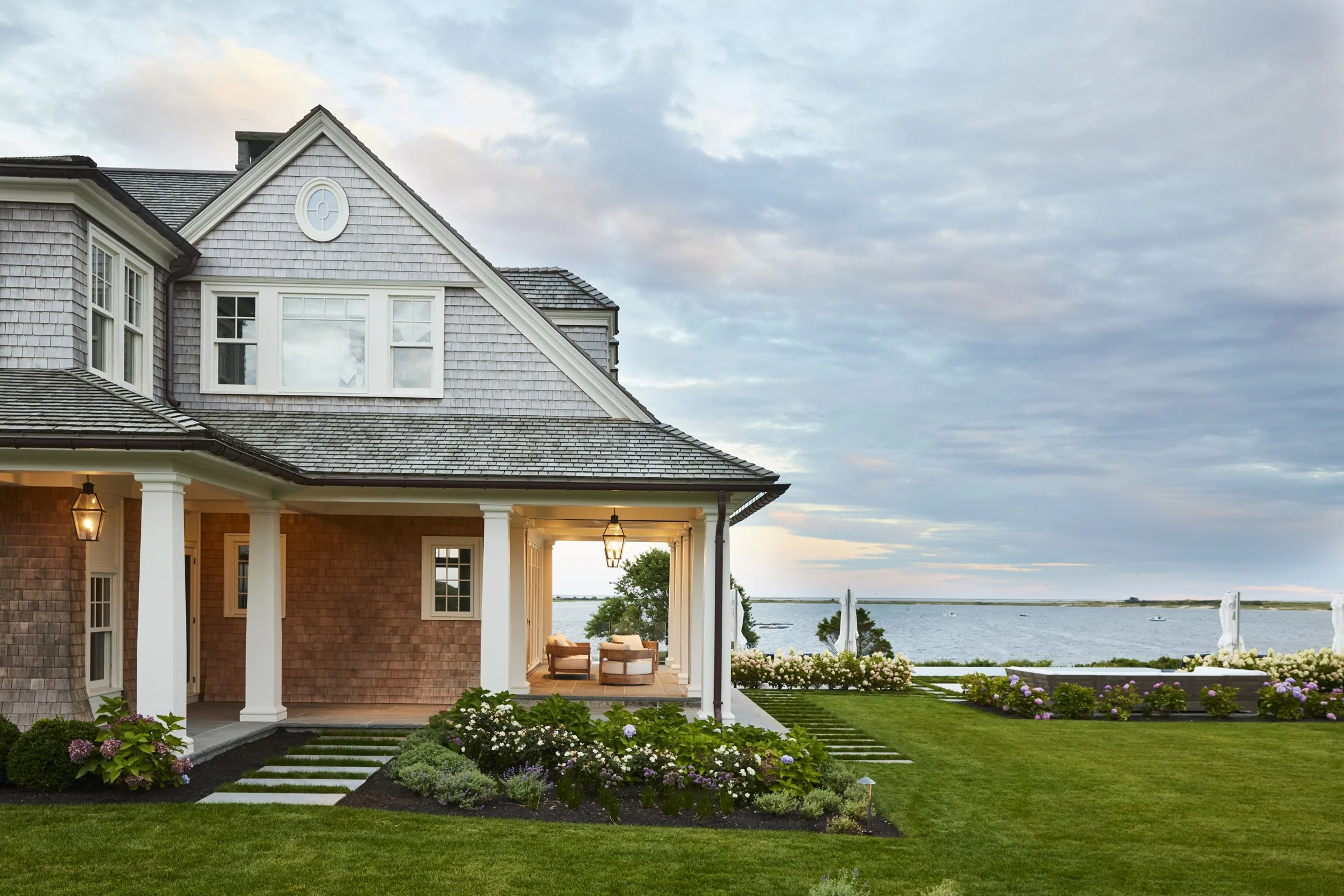 A house with a shingle roof, white trim, and a covered porch overlooking a waterfront with boats, green lawn, and flowering plants, under a partly cloudy sky. Architectural photograph of a Cape Cod coastal home. 