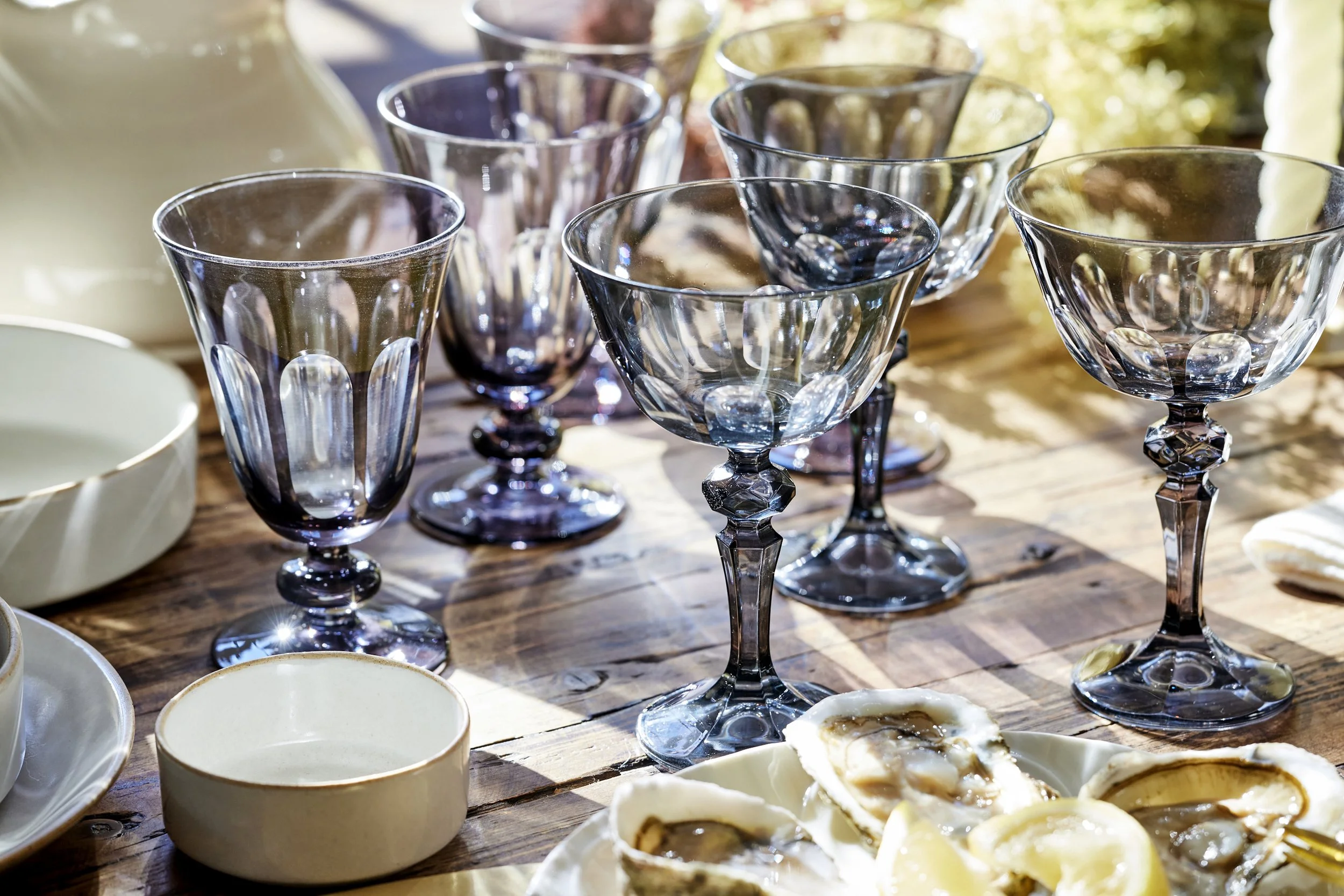 A set of purple and clear glass goblets on a wooden table, with some oysters and empty bowls nearby, in a sunny outdoor setting.