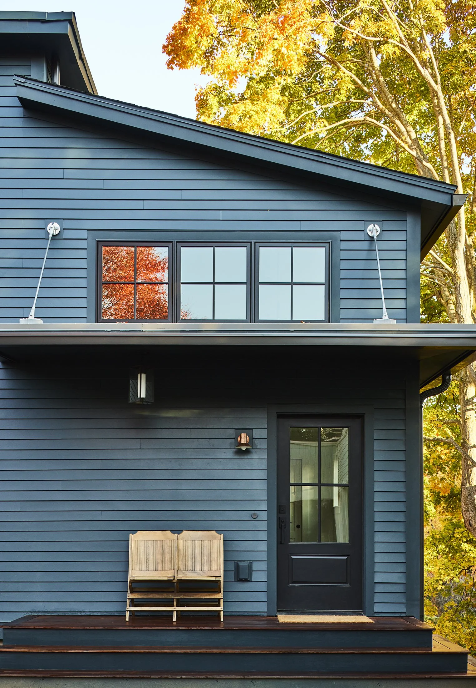 Front of a modern blue house with black window frames, a small porch with wooden steps, a bench, and autumn trees in the background.